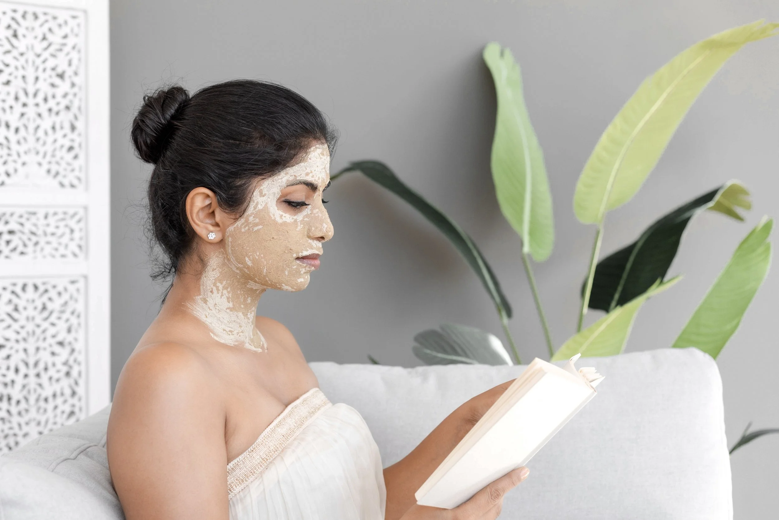 A woman with dark hair tied in a bun, with a cream-colored face mask applied, sitting on a light-colored sofa, reading a book in a room with gray walls and large green houseplants.