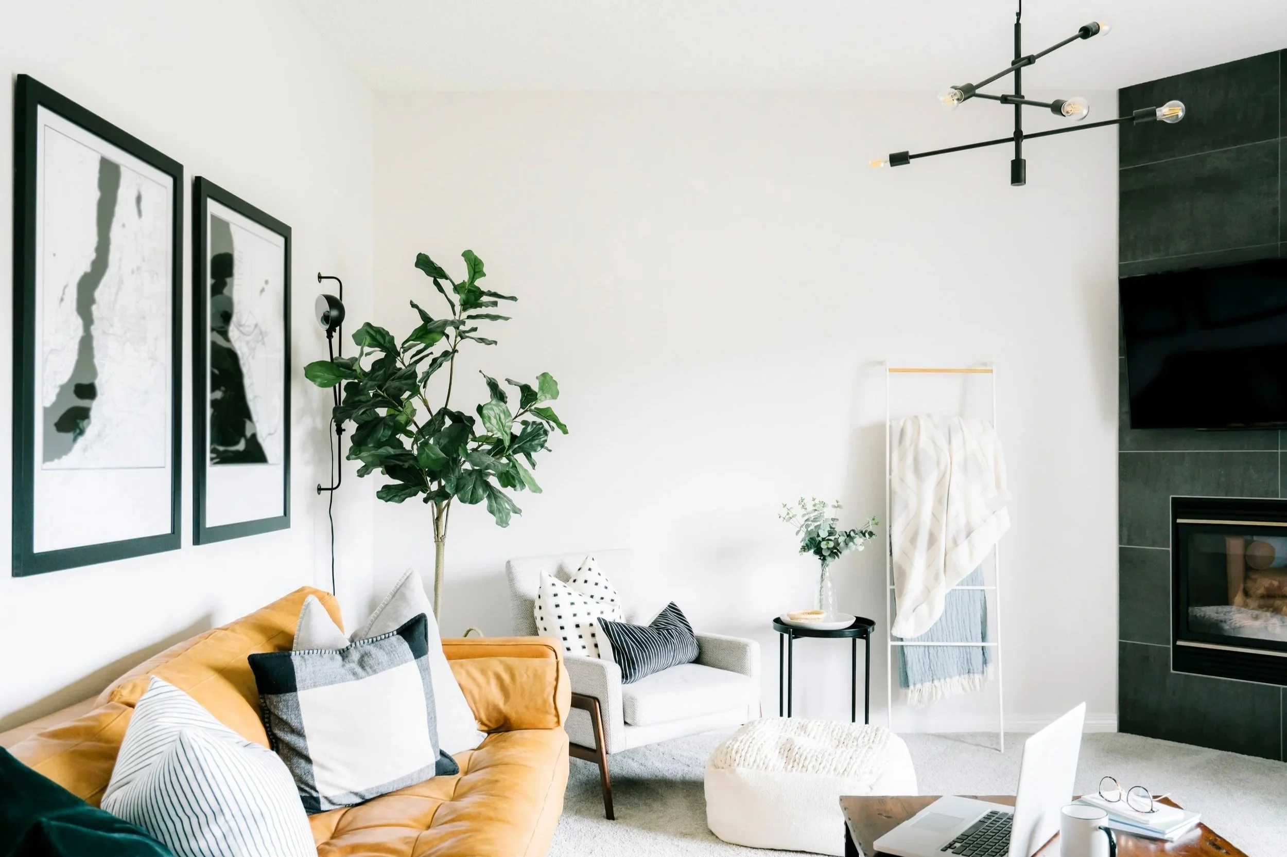 Modern living room with white walls, black framed art, green plants, yellow sofa, patterned pillows, white armchair, small black table, and a TV on the wall.