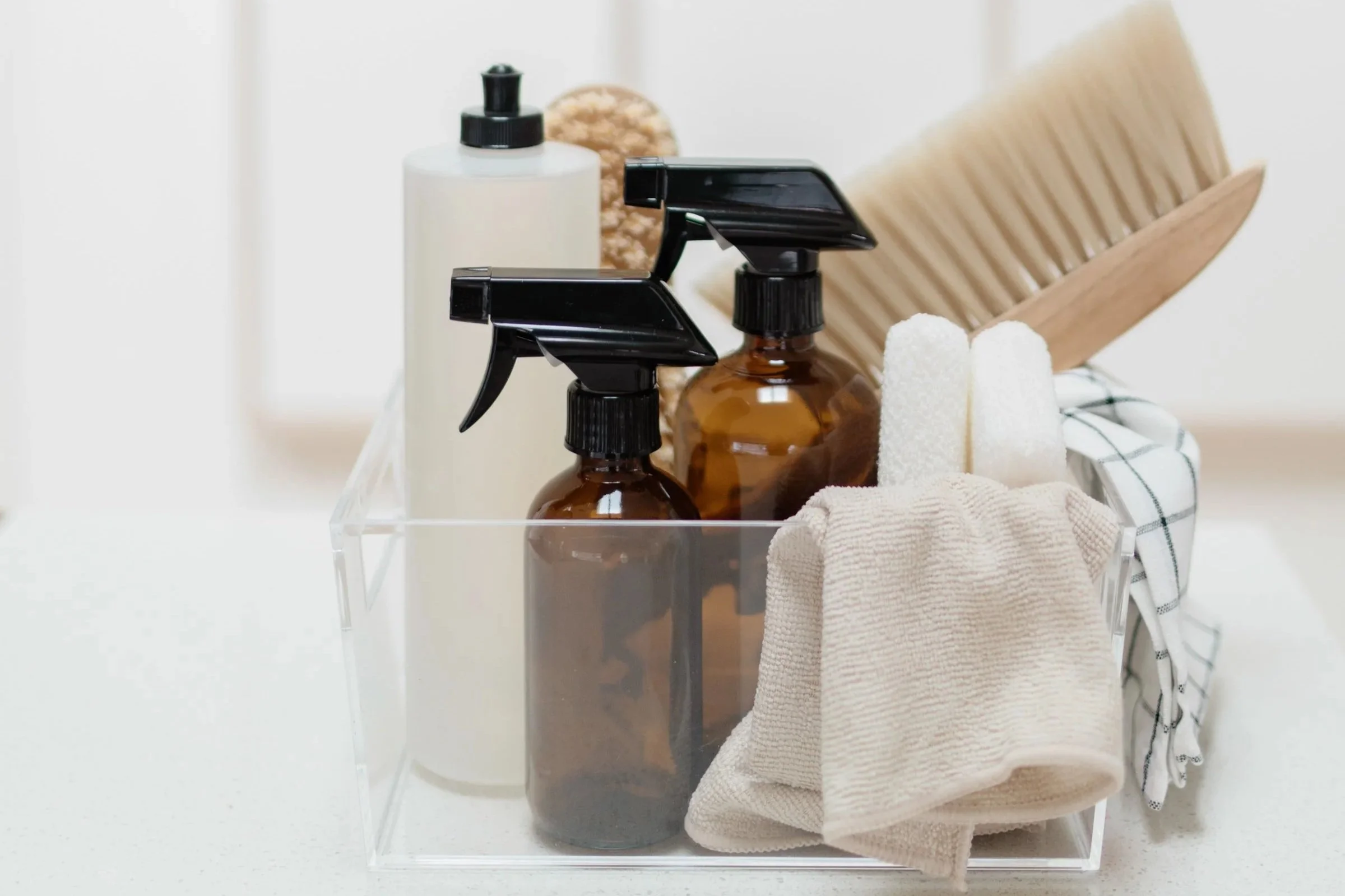 Cleaning supplies organized in a clear container, including spray bottles, towels, and a wooden brush.