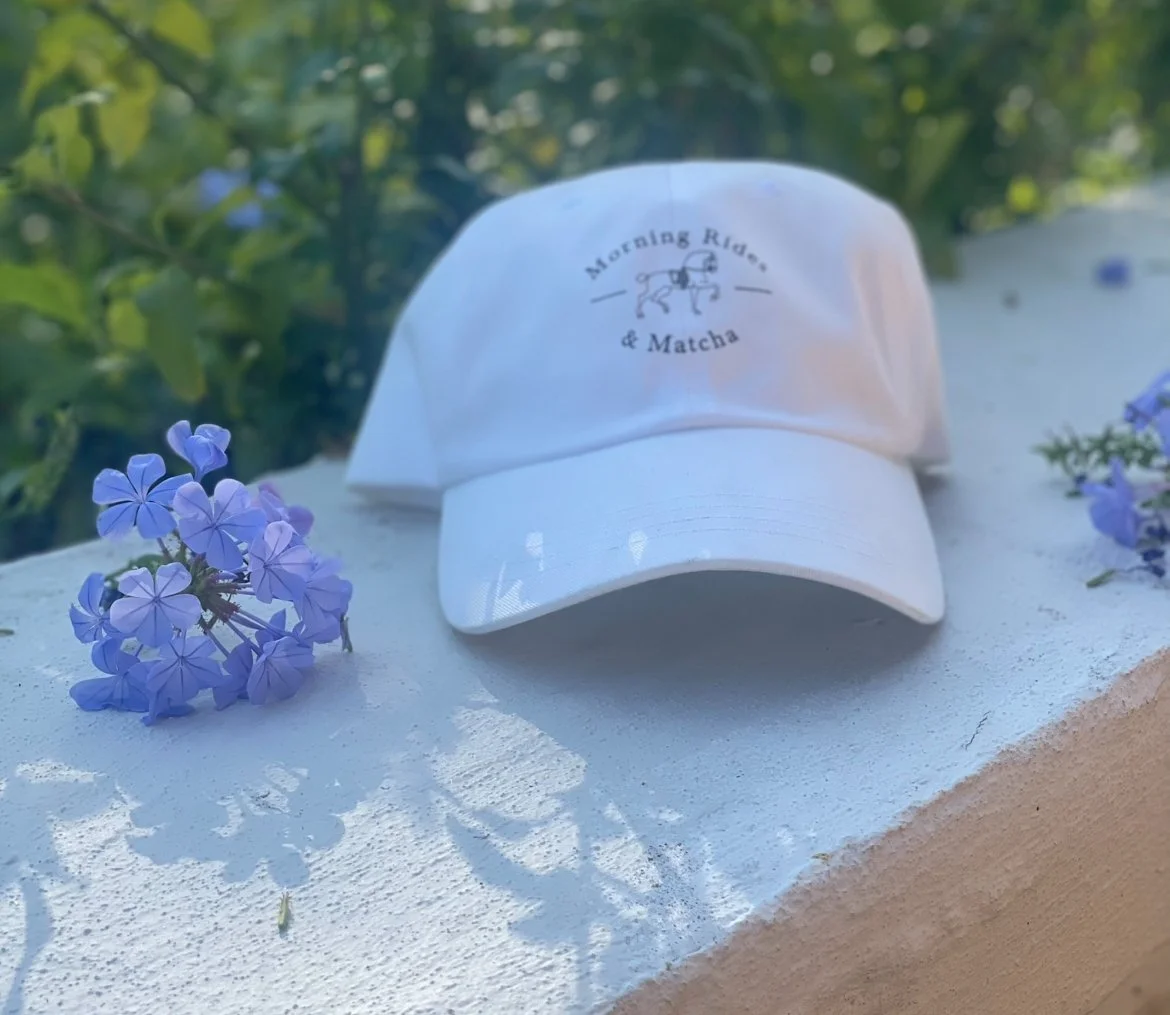White baseball cap with 'Morning Ride & Matcha' logo, placed on a white surface with purple flowers nearby, outdoors with green foliage in the background.