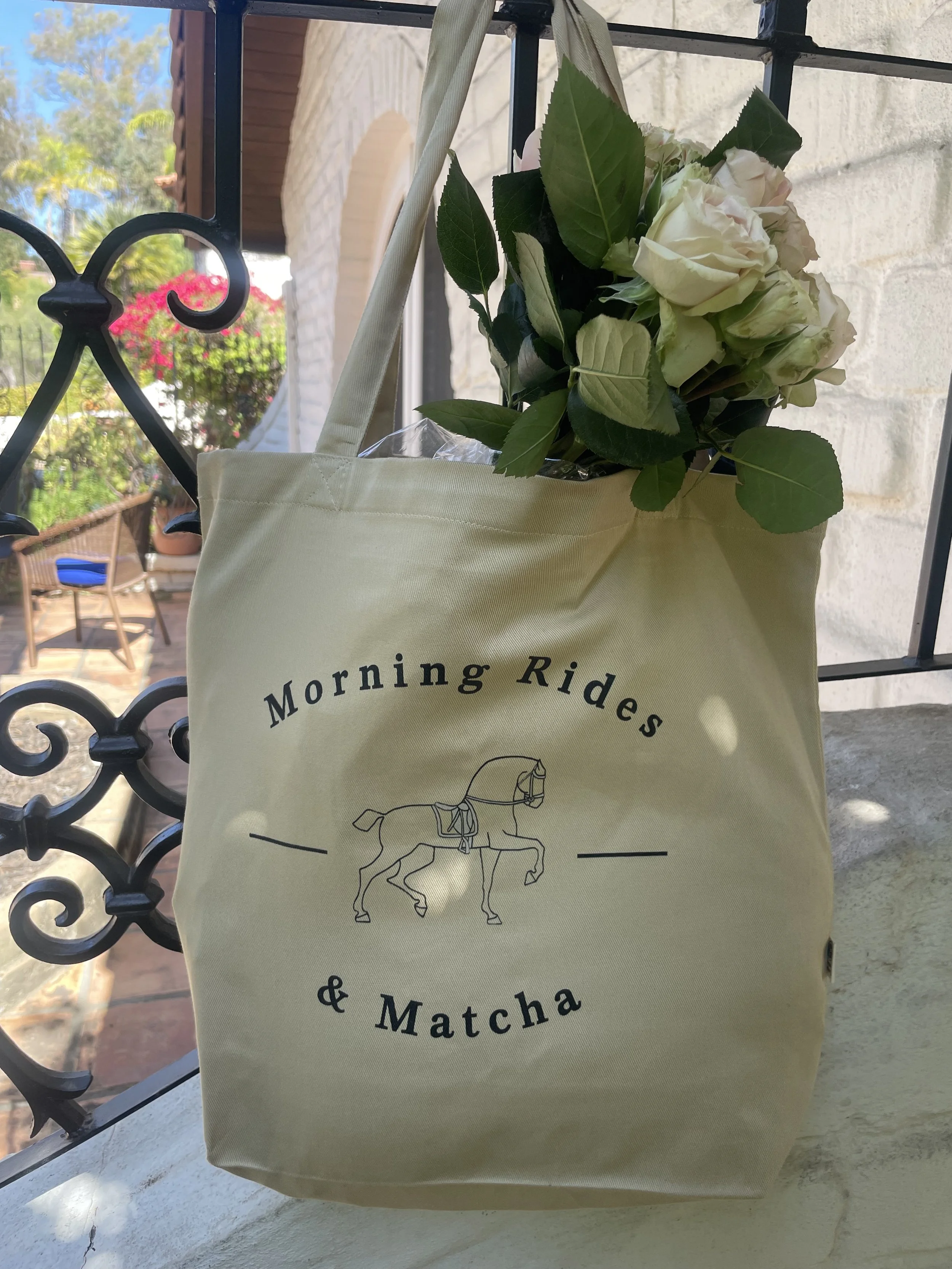 A beige tote bag with the text "Morning Rides & Matcha" and a drawing of a horse in the center, hanging against a railing on a porch, filled with a bouquet of white roses and green leaves.
