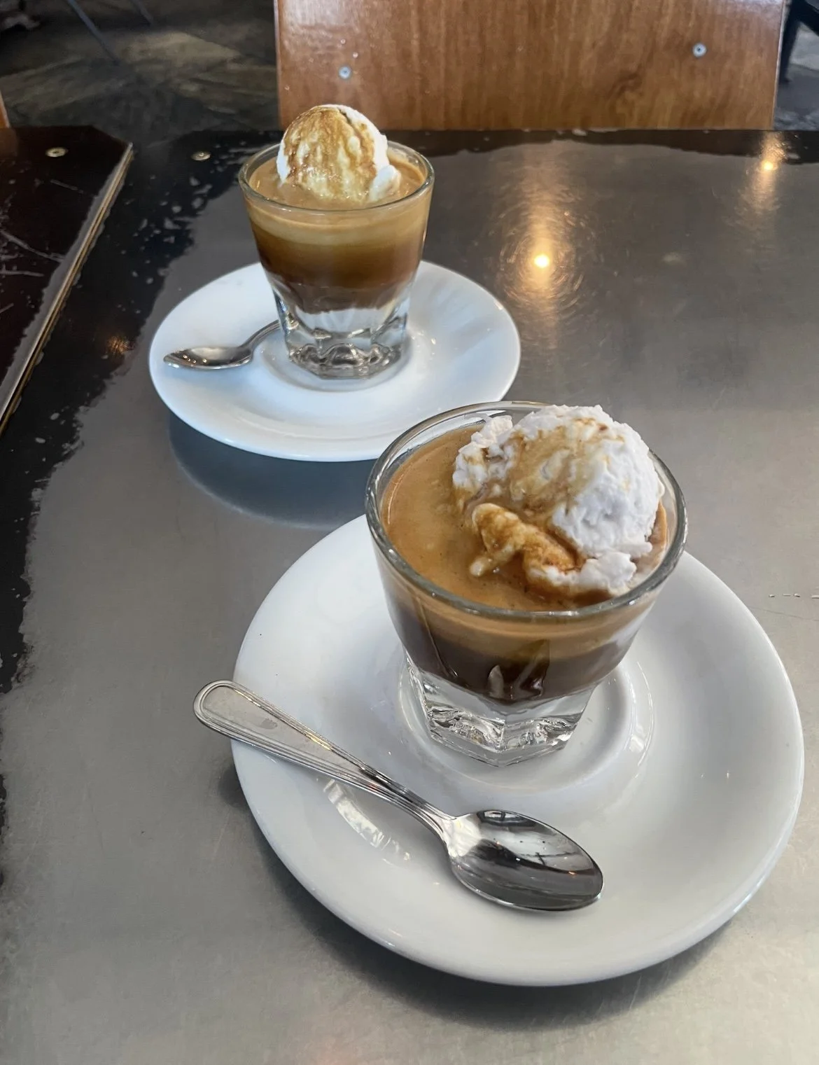Two glasses of chocolate dessert topped with a scoop of vanilla ice cream on a white plate and saucer, on a reflective dark gray table.