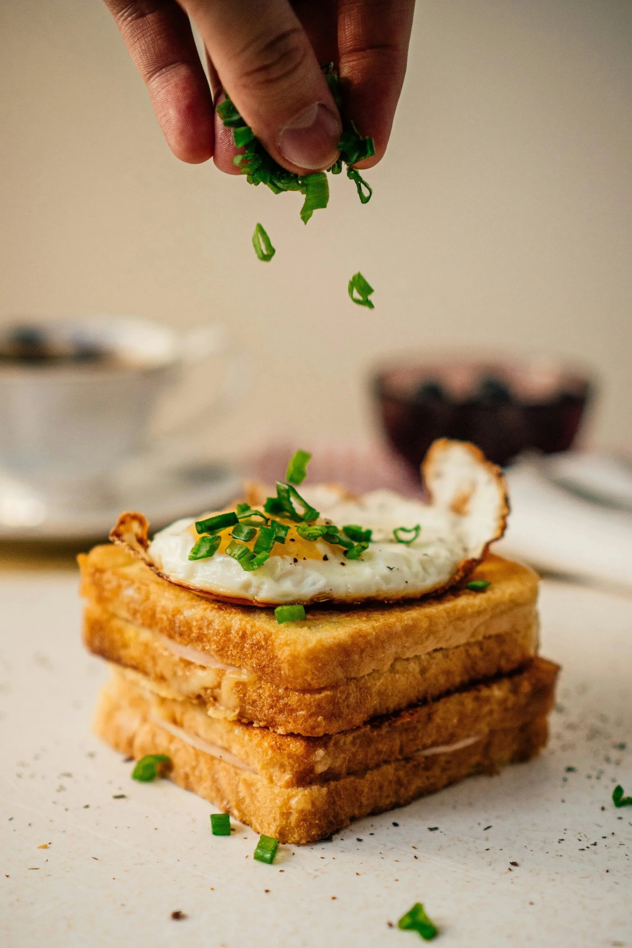 A hand sprinkling chopped green onions onto a stack of toasted bread topped with a fried egg.