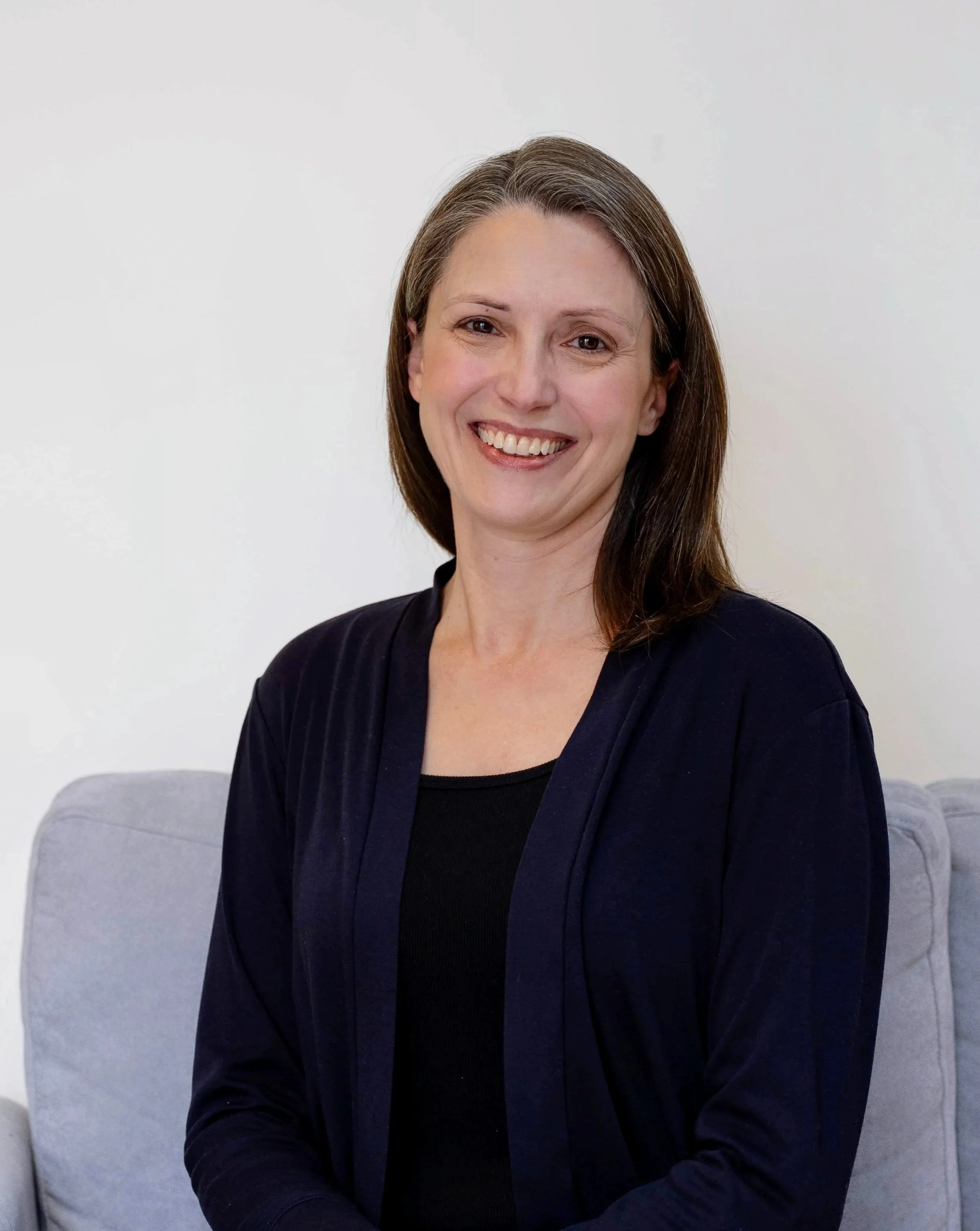Portrait of a woman with brown hair, smiling, wearing a black top and a dark cardigan, sitting on a light gray couch against a white wall.