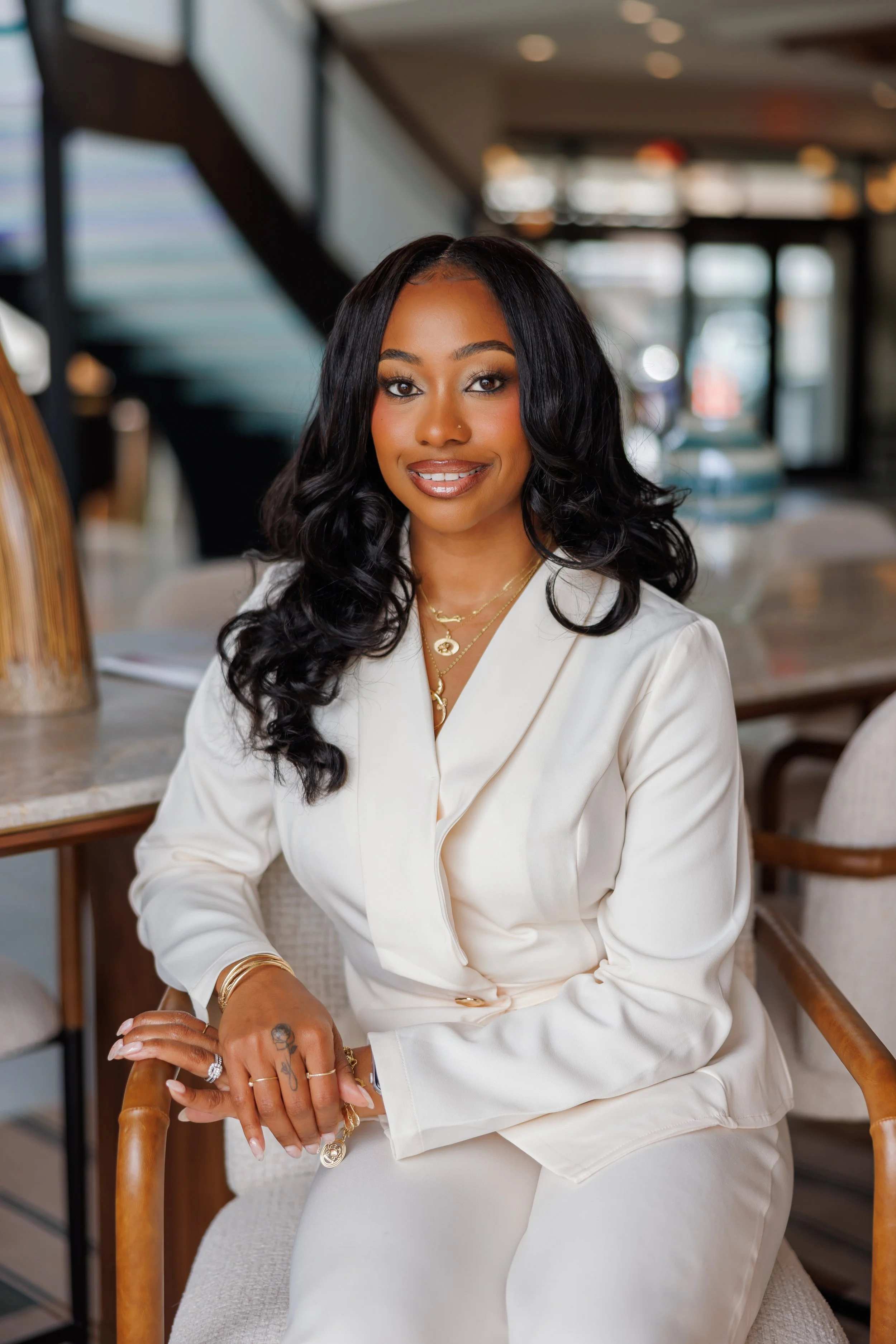 A woman with long, curly black hair, wearing a white blazer and jewelry, sitting on a chair in a modern, well-lit indoor space.