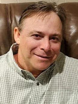 A man with short brown hair and a fair complexion, wearing a checkered button-up shirt, smiling while sitting on a brown leather chair.