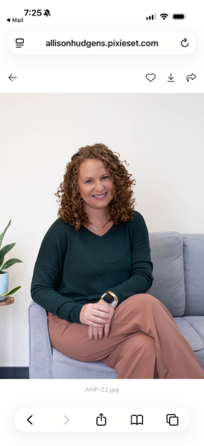A woman with curly red hair sitting on a gray couch, wearing a dark green sweater and beige pants, smiling at the camera.