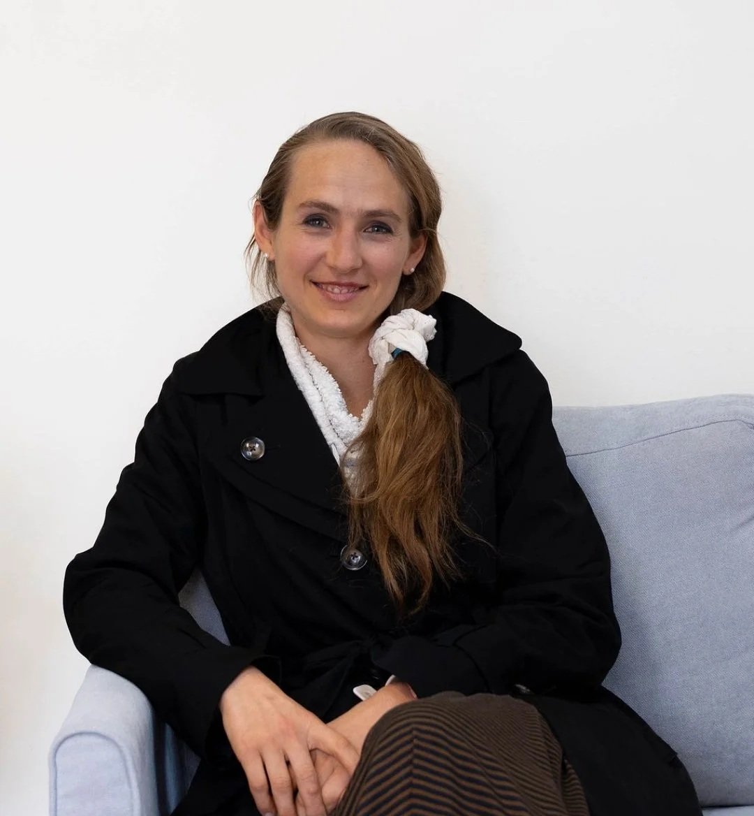 A woman with long red hair tied in a ponytail, wearing a black coat, sitting on a light blue sofa, smiling at the camera.