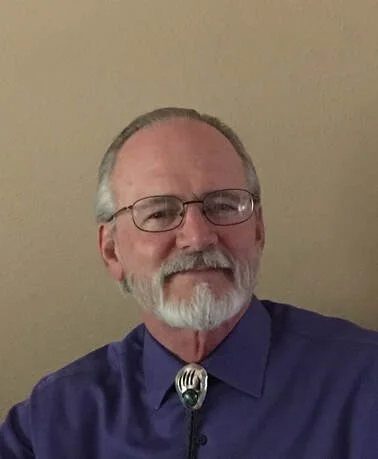 A man with glasses, a beard, and a mustache wearing a blue shirt and a bolo tie, smiling at the camera.