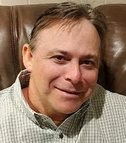 A middle-aged man with light brown hair and a slight smile, wearing a checkered shirt, sitting on a brown leather chair.