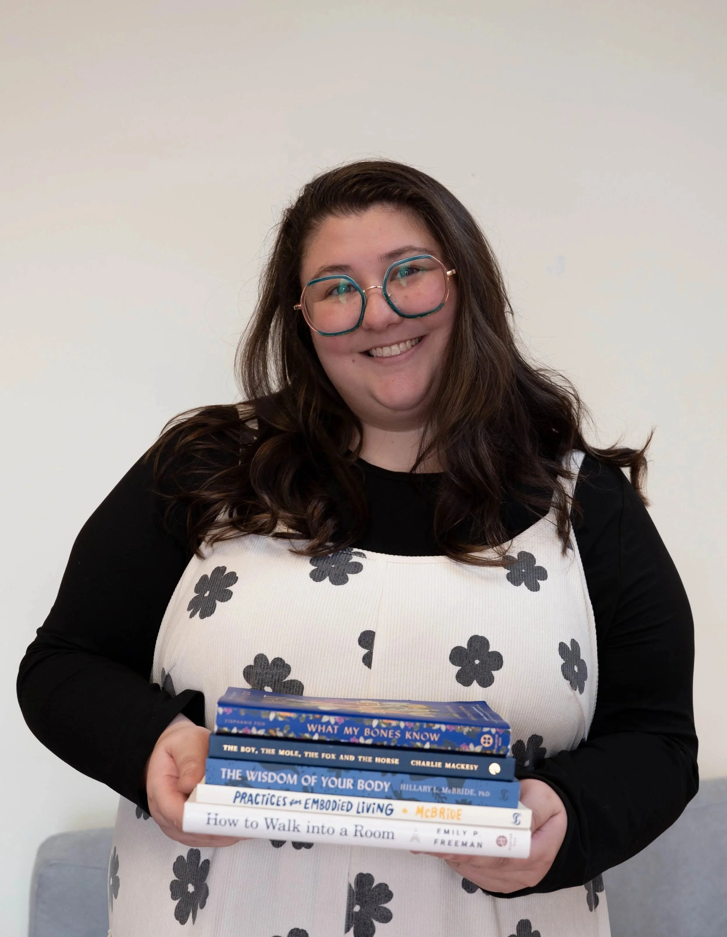 A woman with long brown hair, glasses, and a black shirt, holding a stack of books and smiling at the camera against a plain, light-colored background.