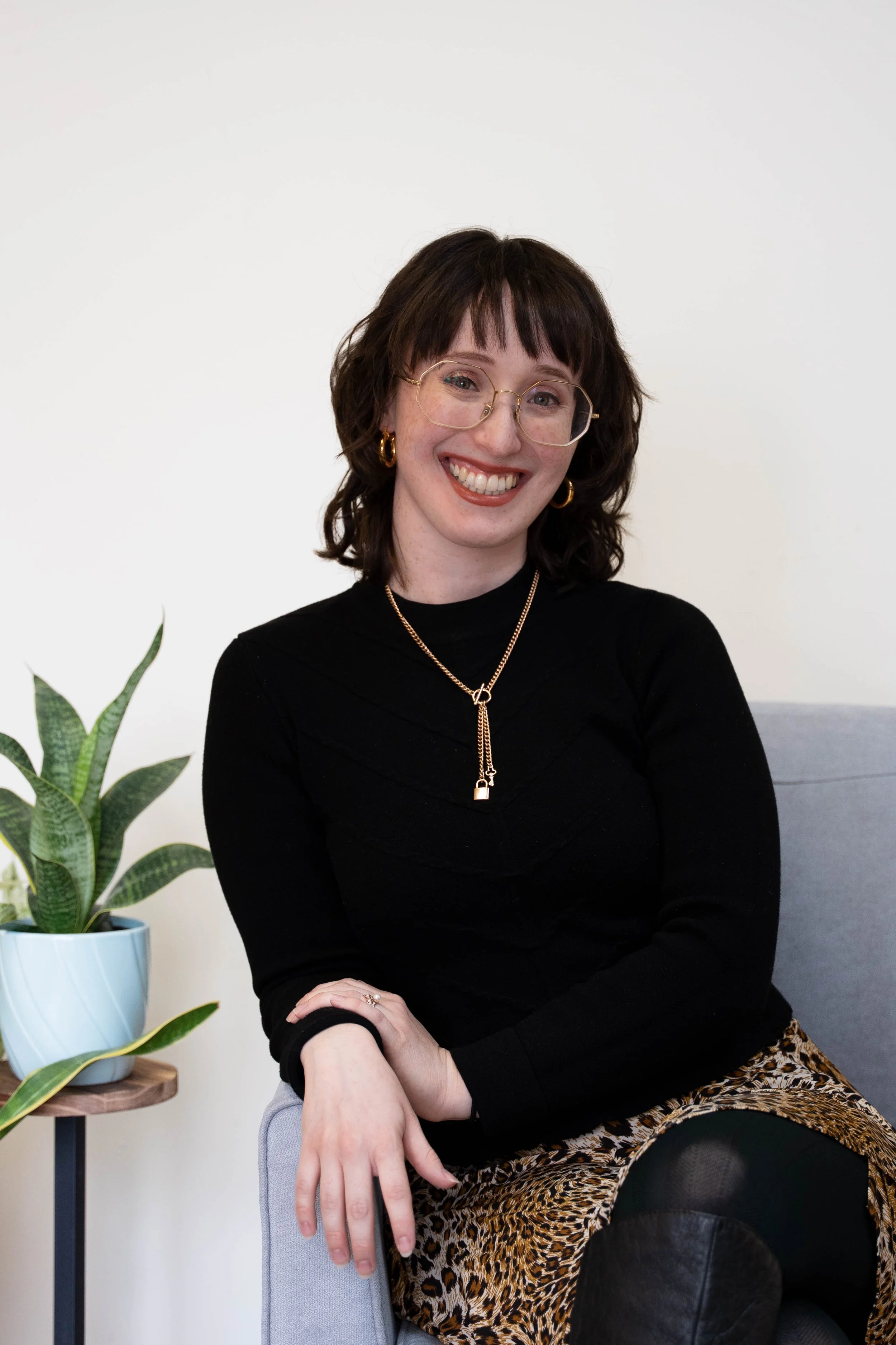 A woman with dark brown hair, glasses, and gold jewelry, sitting on a chair next to a potted snake plant, smiling at the camera.