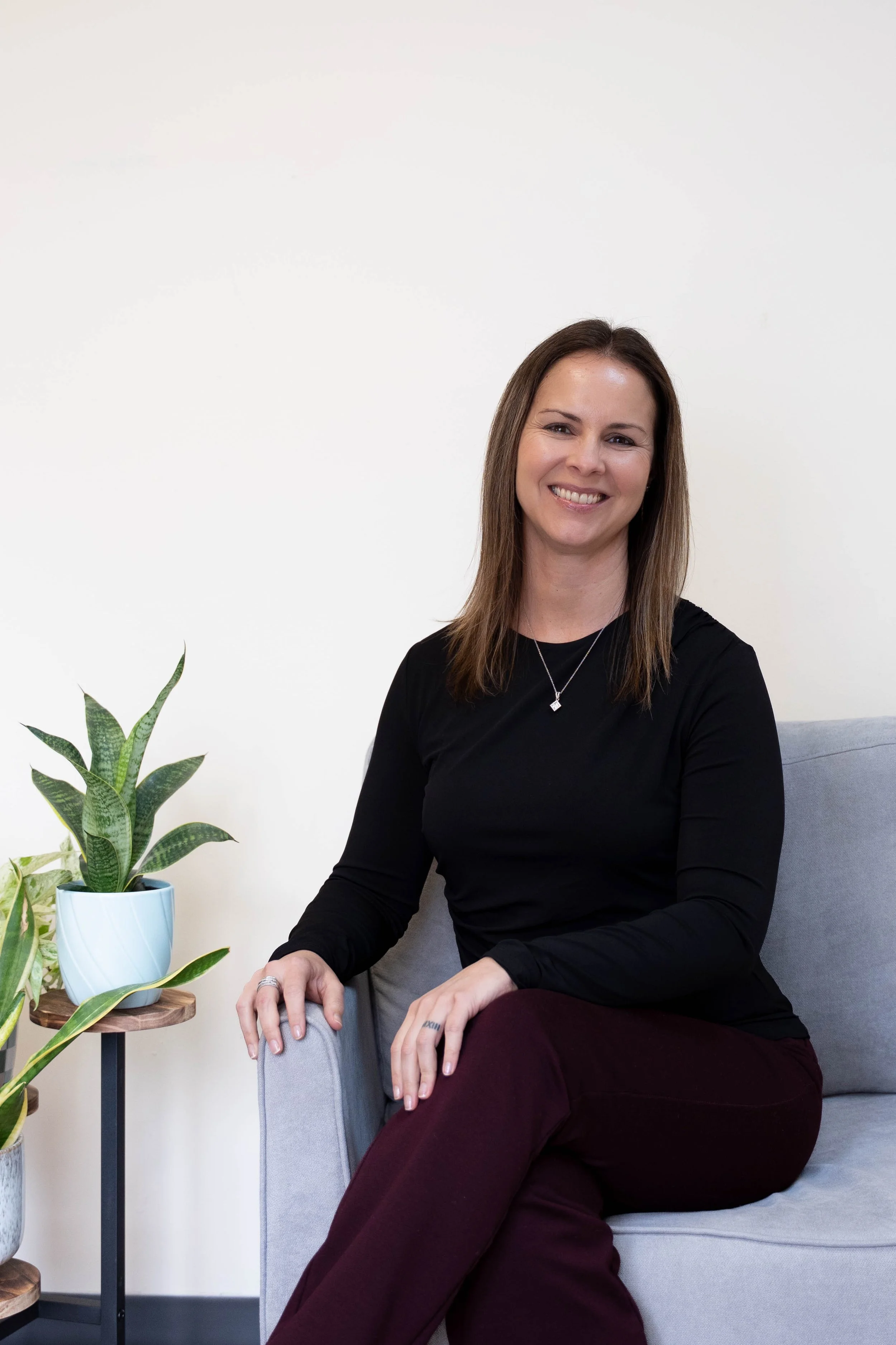 A woman with shoulder-length brown hair sitting on a light gray sofa, smiling, wearing a black long-sleeve shirt and maroon pants, with a necklace, in a room with plants on a side table.