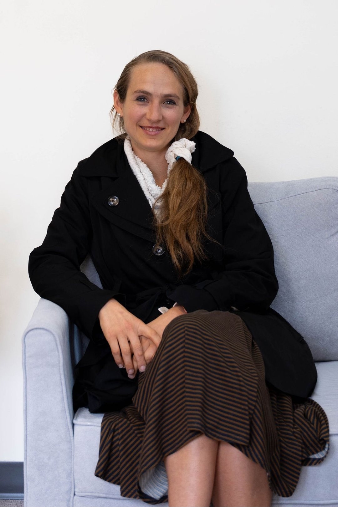 A woman with long, reddish-brown hair in a side ponytail, sitting on a light gray sofa, wearing a black trench coat and a striped skirt, smiling at the camera.