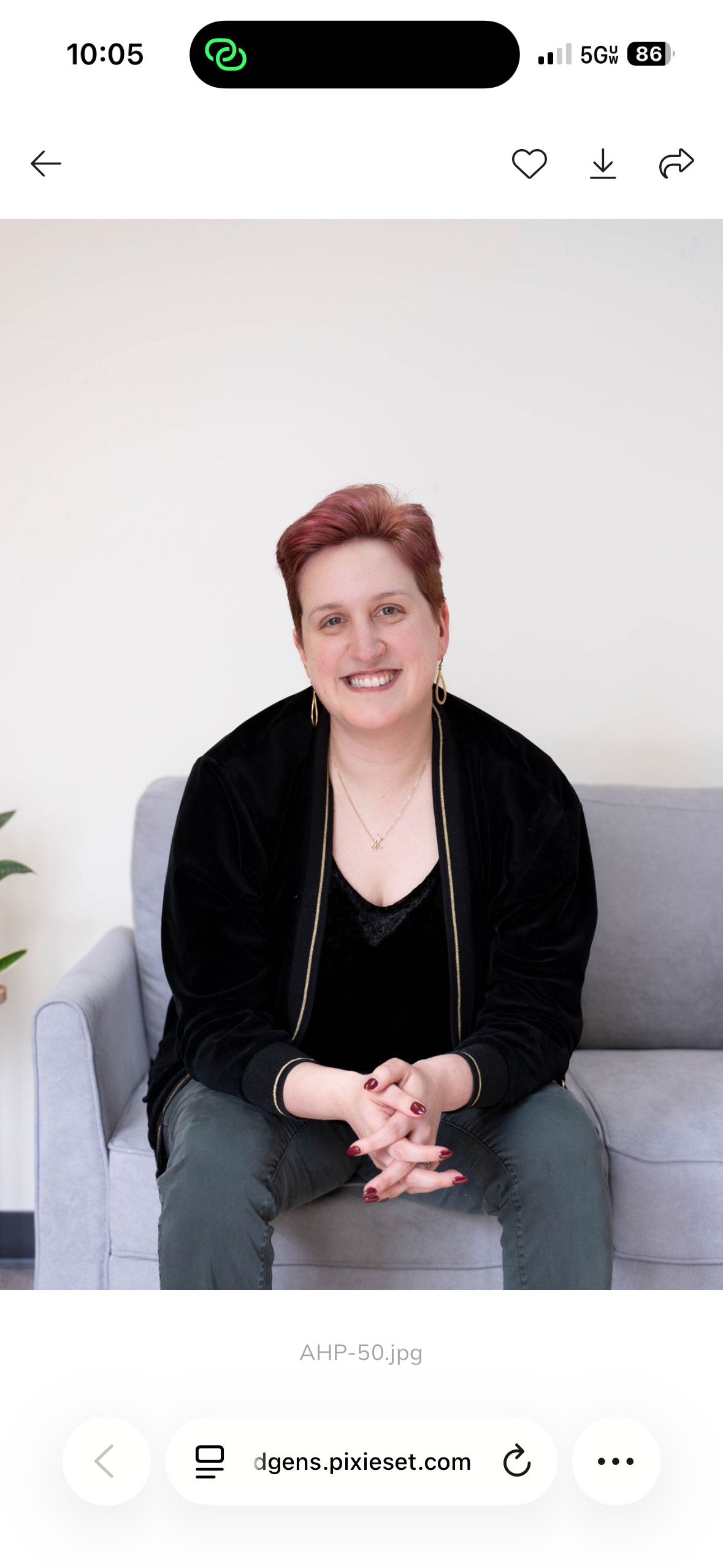A woman with short red hair, wearing a black jacket, black top, and gray pants, is sitting on a light gray sofa with her hands clasped. She is smiling and looking at the camera, with a white wall background and a small plant partially visible on the left.