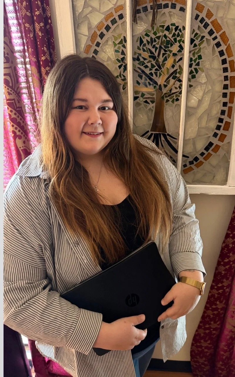 A young woman with long brown hair, wearing a black top and a striped button-up shirt, is holding a closed black HP laptop. She is standing near a decorative stained glass window featuring a tree design, with purple and red patterned curtains visible nearby.