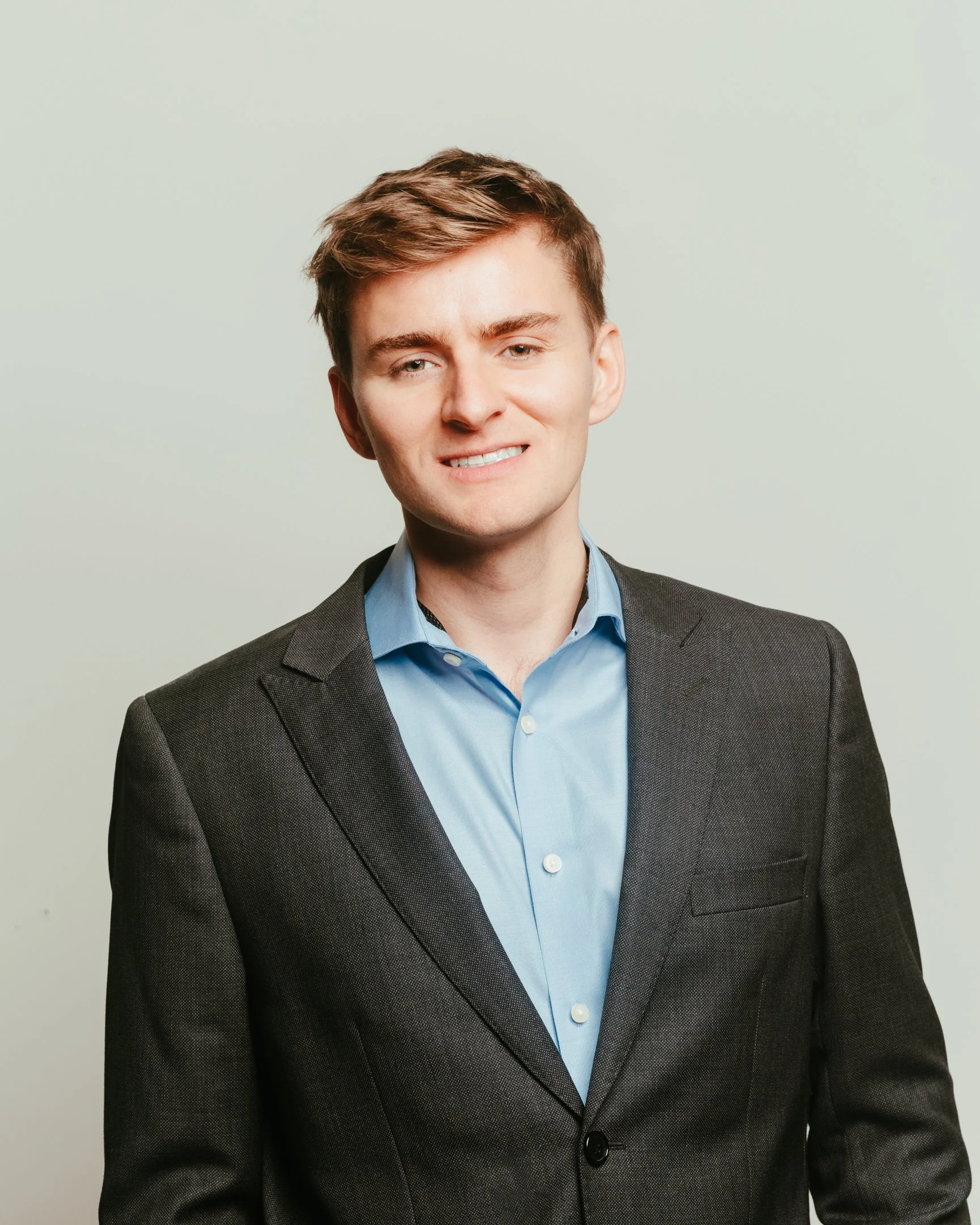 A young man with brown hair, wearing a dark gray blazer and blue shirt, standing against a light gray background.