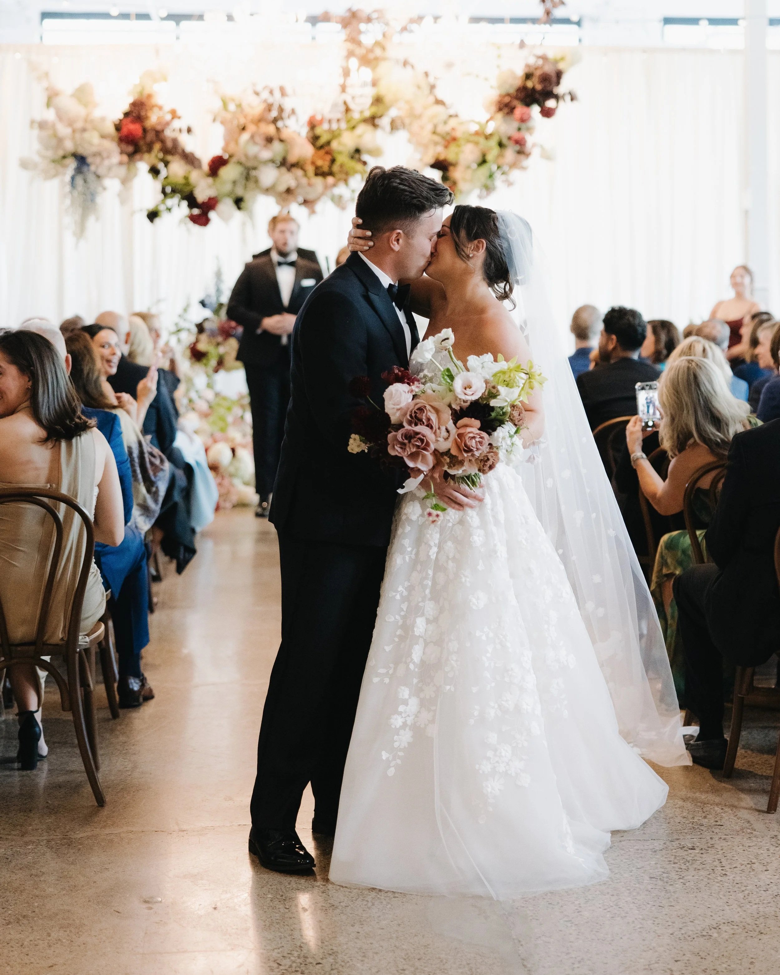 A bride and groom kiss during their wedding ceremony in a decorated indoor venue.