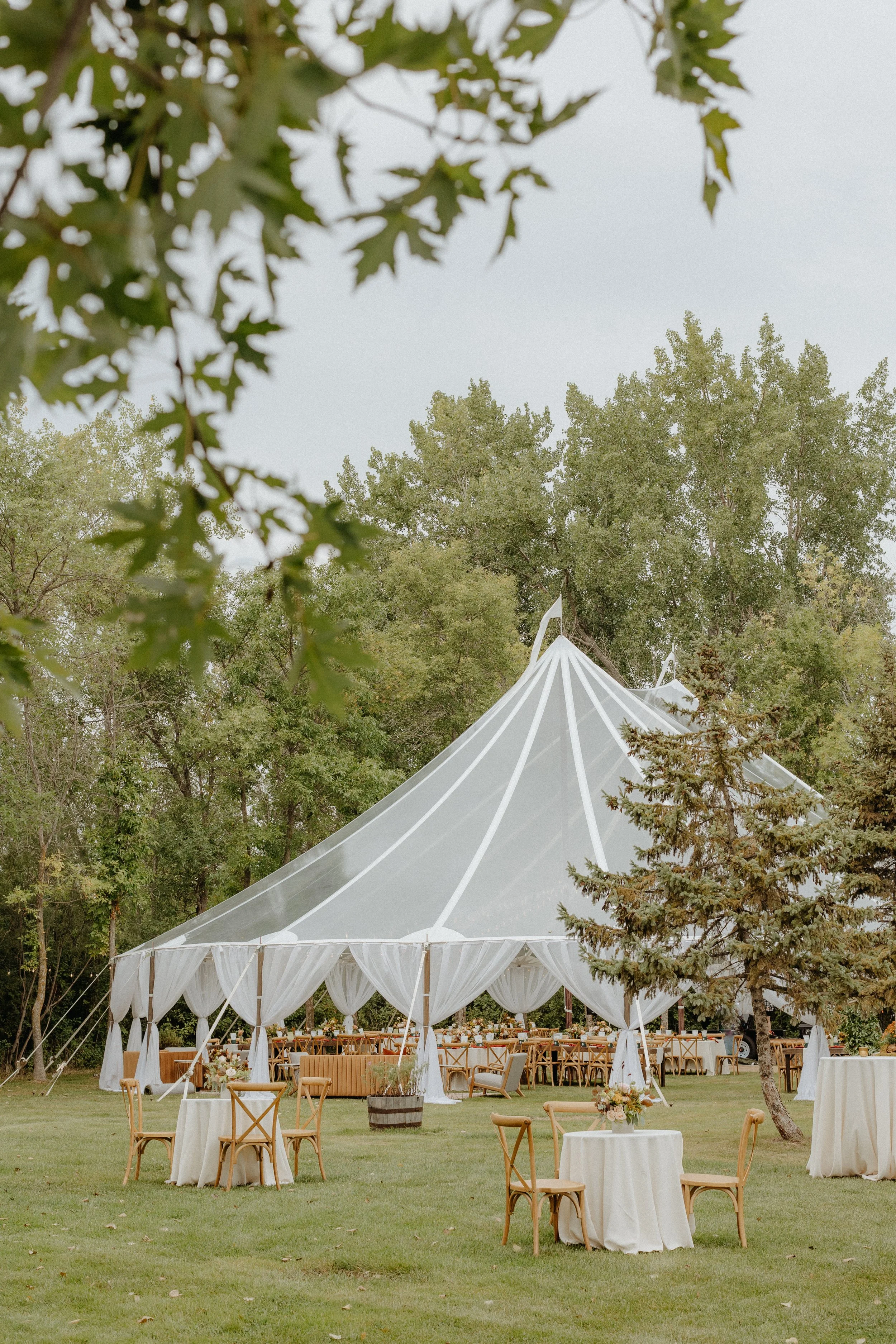 Outdoor wedding reception setup with a large white tent, tables with white tablecloths, chairs, and floral centerpieces, surrounded by trees.