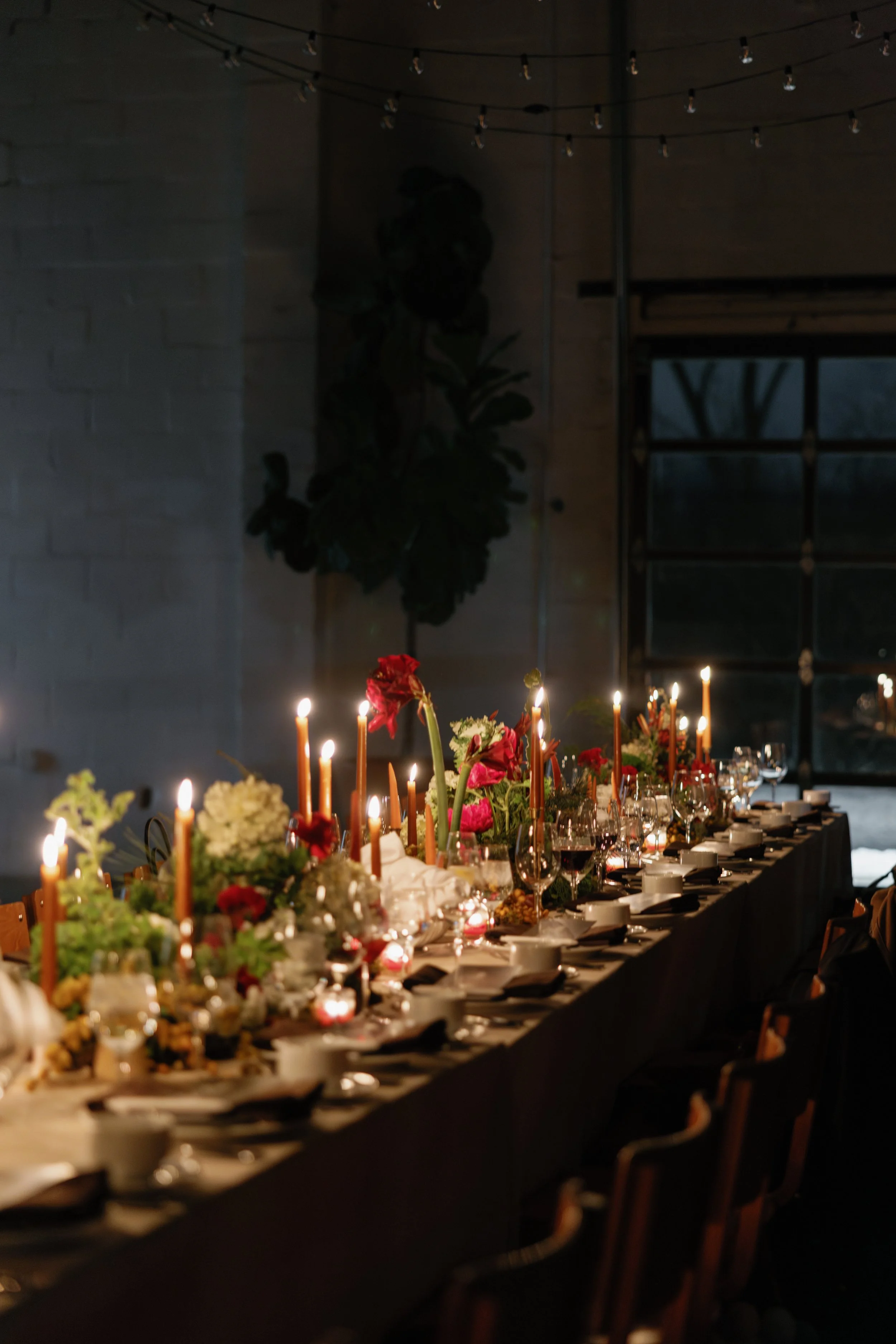 A long dining table decorated with flowers, candles, and tableware in a dimly lit indoor setting.