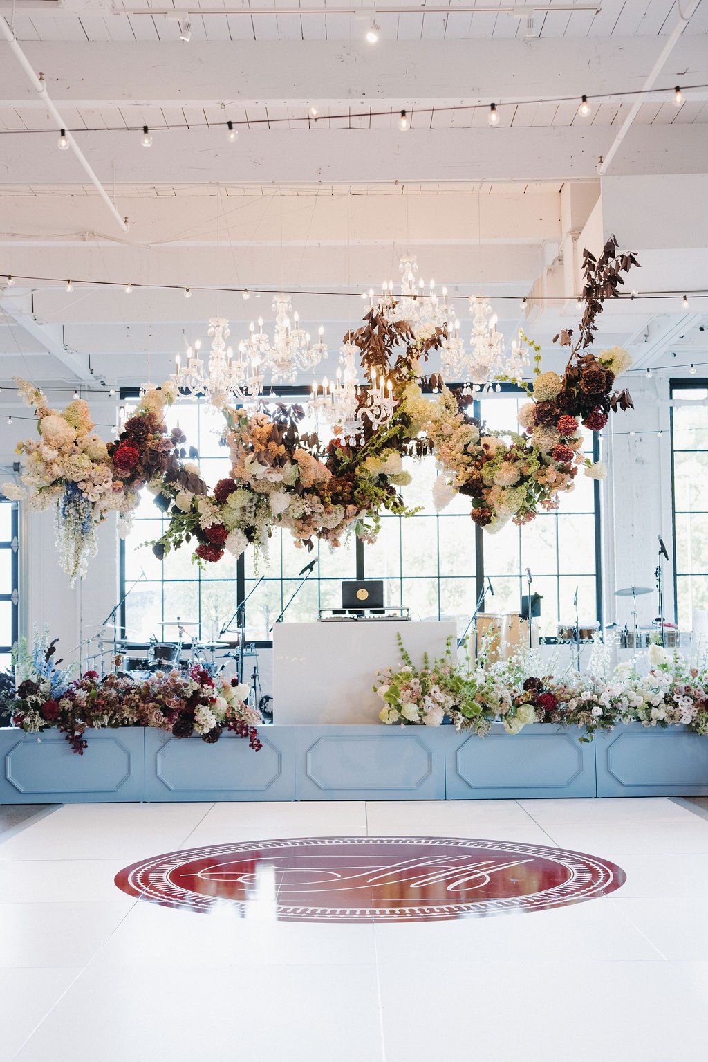 Elegant event space decorated with large floral arrangements hanging from the ceiling, chandeliers, and a decorated floor with a monogrammed design in the center. Microphones and musical instruments are set up in the background.