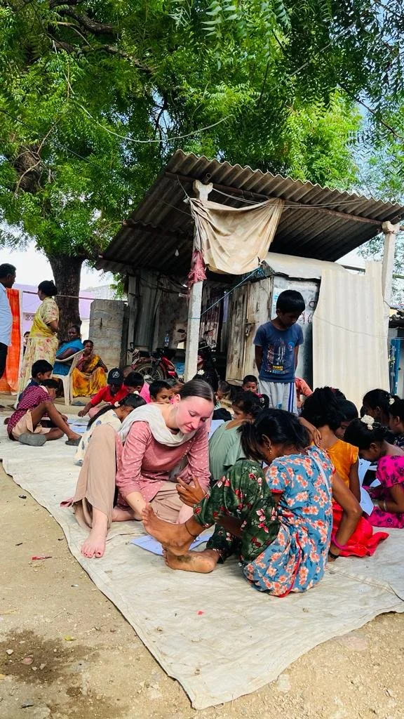A group of people, including children and adults, sitting on a large mat outdoors under a tree near a small makeshift shelter, with a woman interacting with a child in the foreground.