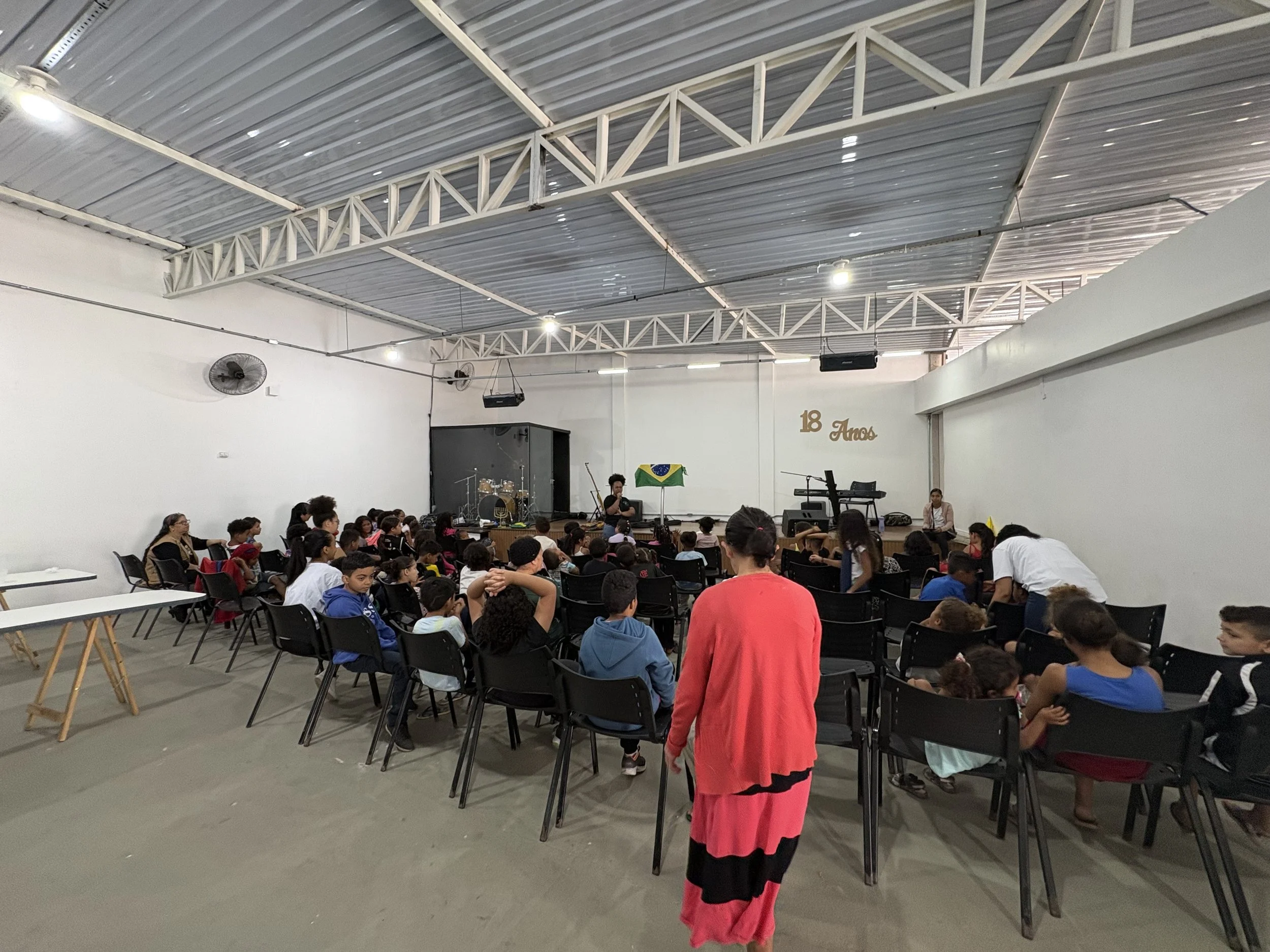 Children and adults seated in chairs watching a performance on a stage with musical instruments and a Brazilian flag in the background, in a large indoor hall decorated for an 18th anniversary celebration.