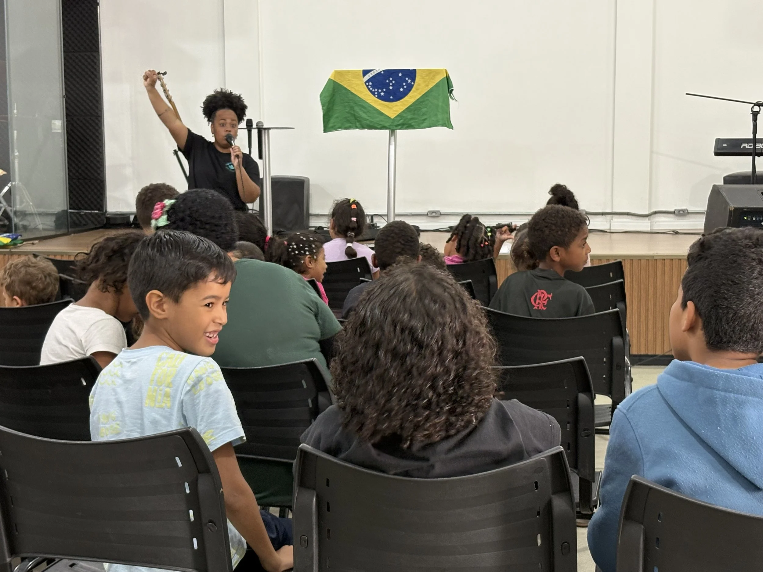 A woman speaking into a microphone on a stage with a Brazilian flag in the background, an audience of children and adults seated facing the stage, some children smiling and listening attentively.