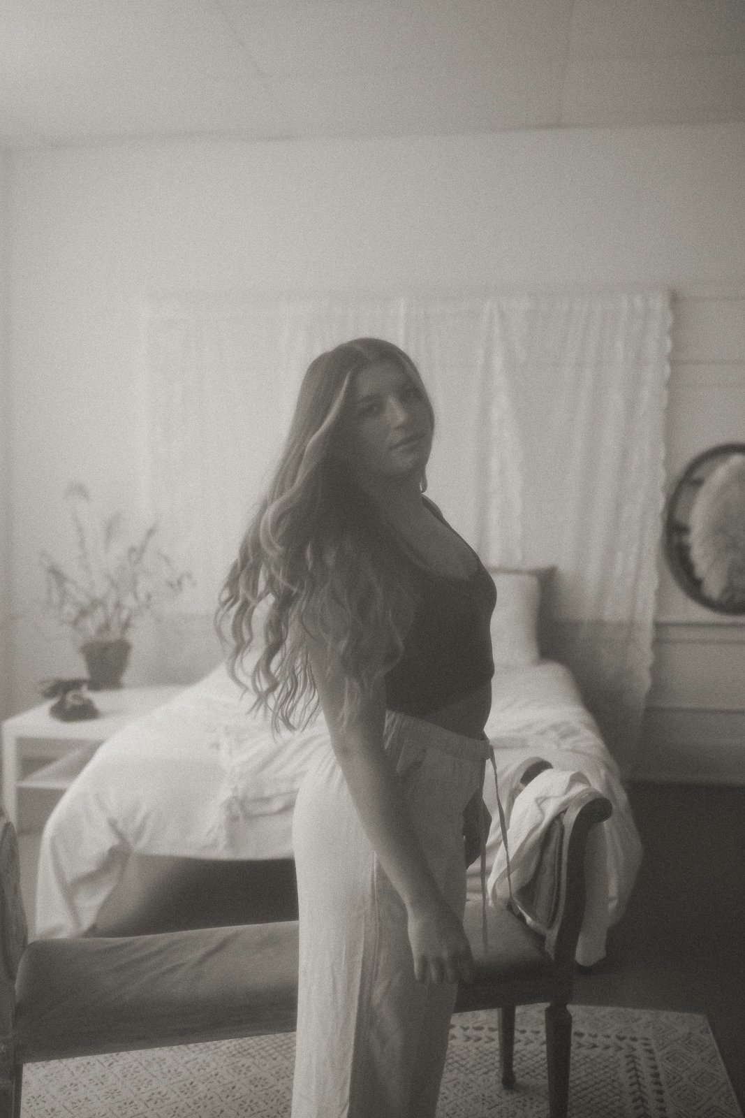 A woman with long, wavy hair standing in a bedroom, wearing a dark top and light-colored pants, looking at the camera.