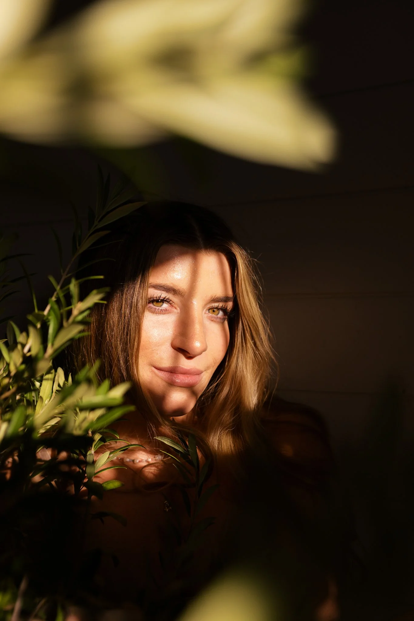A woman with long blonde hair and light eyes peeks through green foliage, with warm sunlight illuminating her face against a dark background.