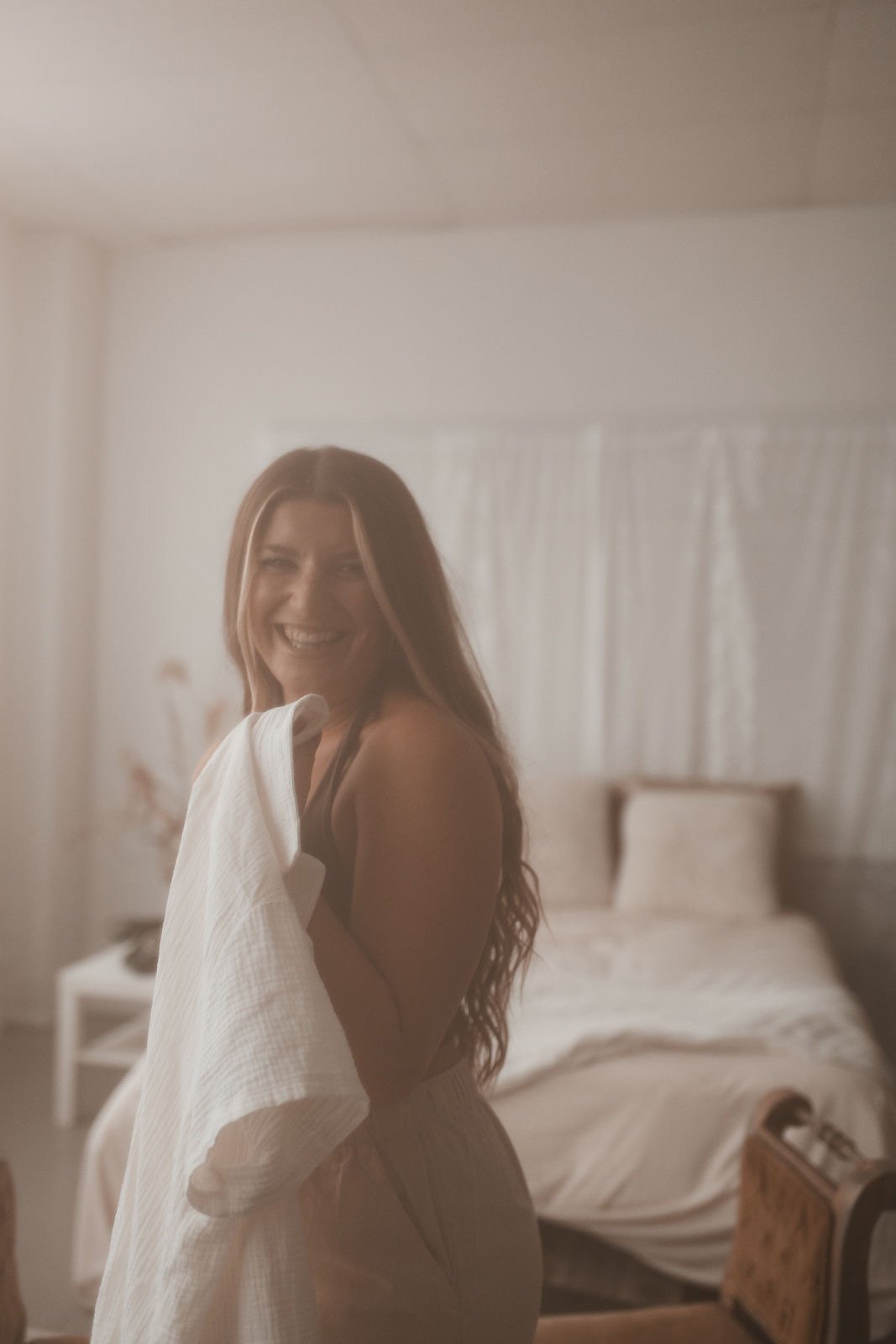 A woman with long reddish-brown hair smiling while holding a white towel in a bedroom with a bed and white curtains in the background.