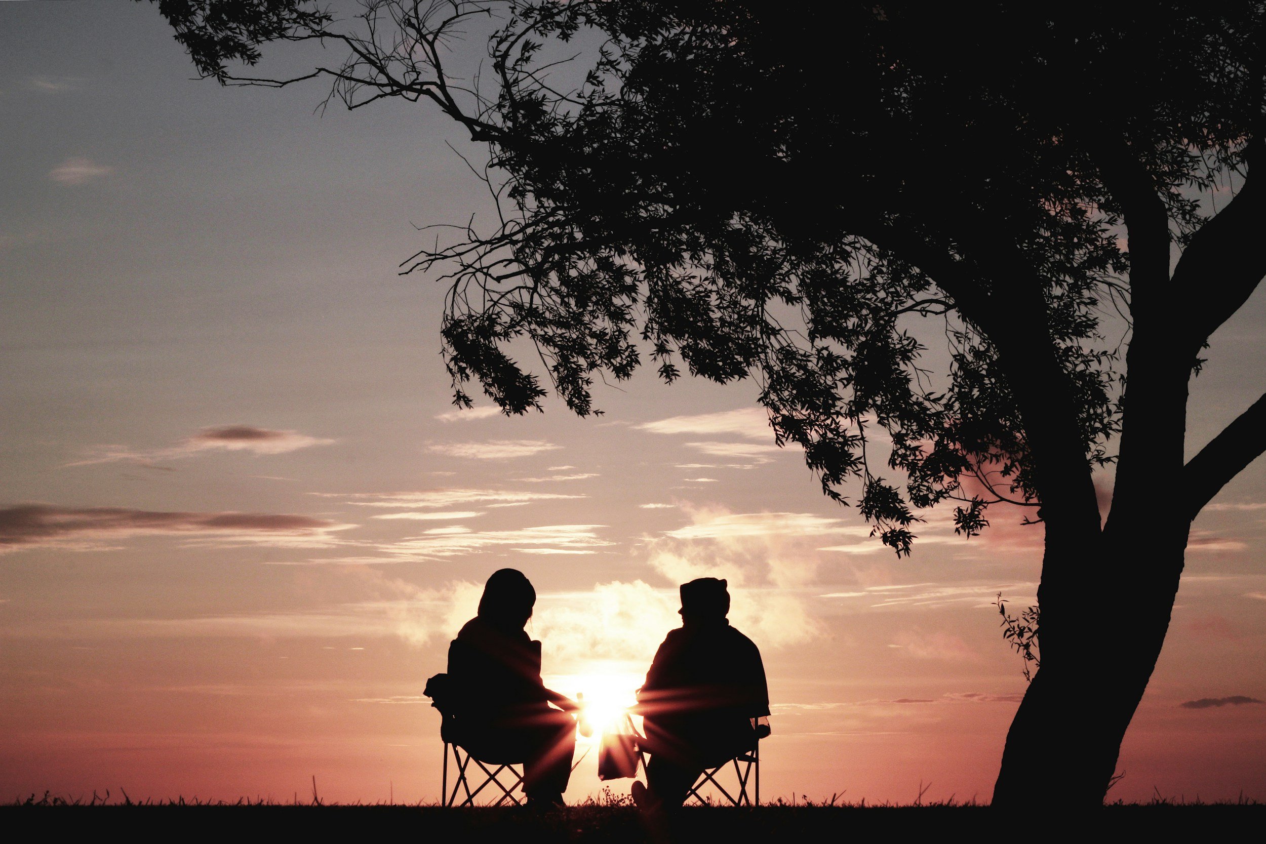 Silhouettes of two people sitting on chairs outdoors at sunset, with a tree on the right and a colorful sky with clouds in the background.