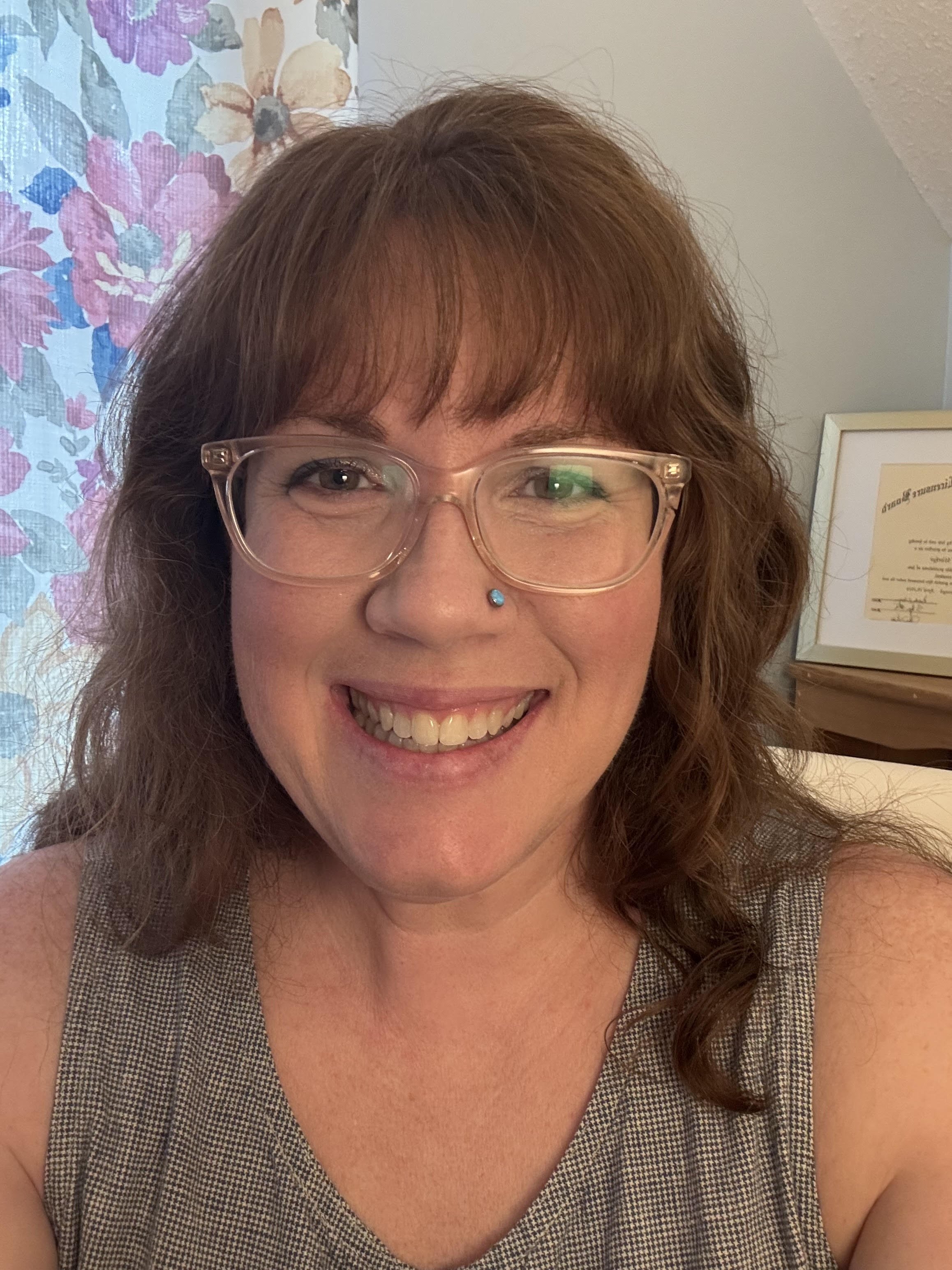 A woman with curly brown hair, wearing glasses with clear frames, smiling, with a blue nose piercing. She is wearing a sleeveless checkered top. In the background, there is a floral curtain and a framed certificate or diploma on the wall.