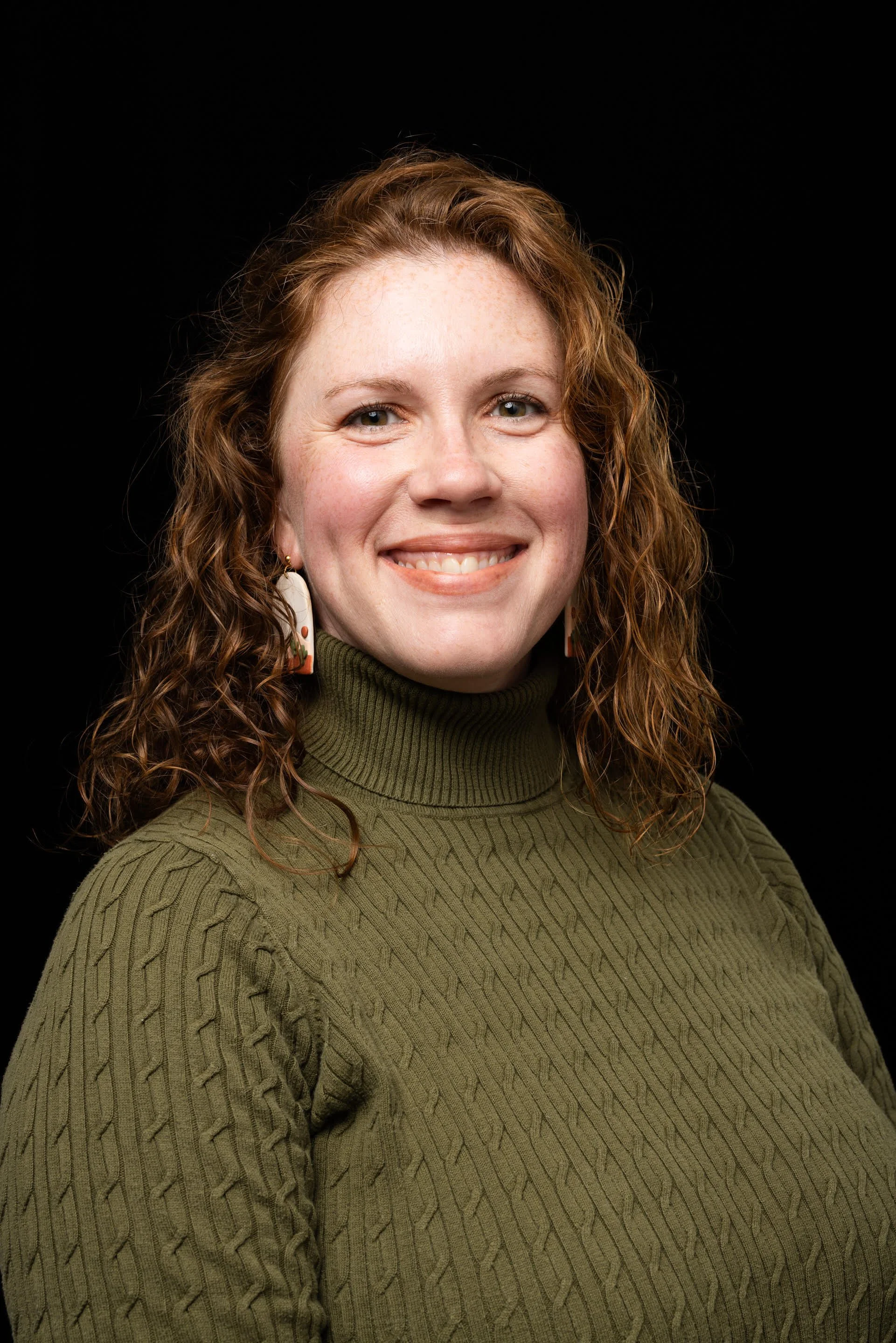 Portrait of a woman with curly red hair, smiling, wearing a green cable-knit turtleneck sweater and dangly earrings, against a black background.