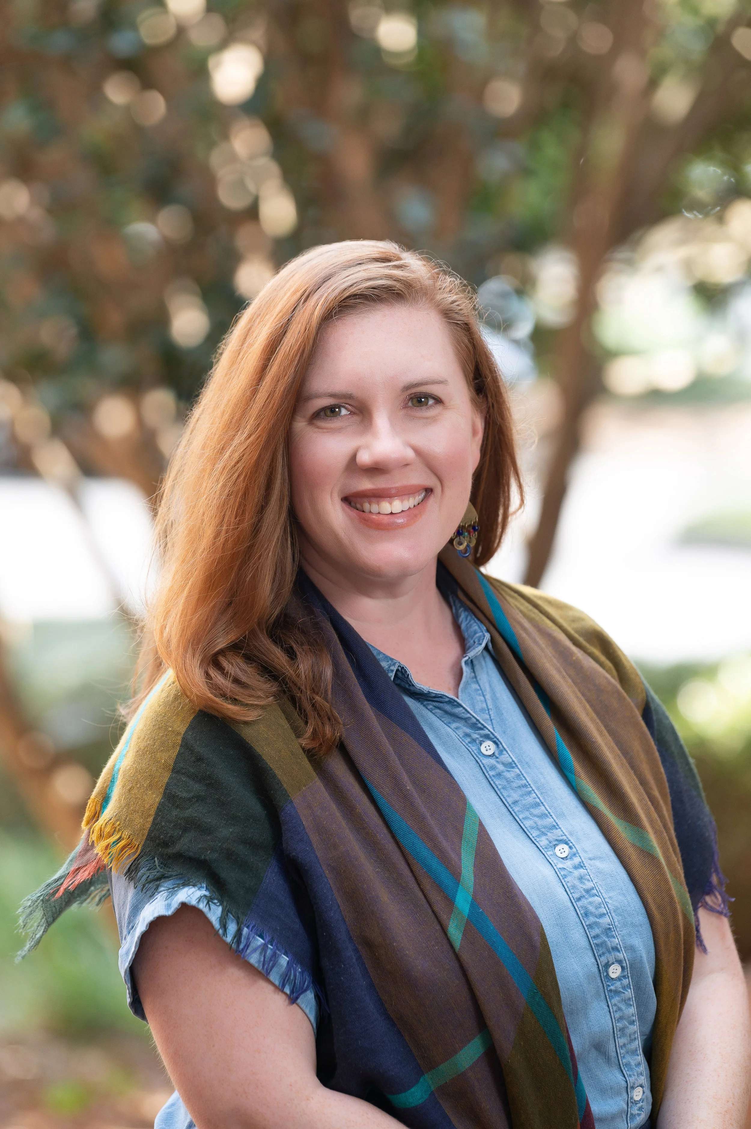A woman with long red hair smiling outdoors, wearing a blue denim shirt and a multicolored plaid scarf, with a blurred background of trees and sunlight.