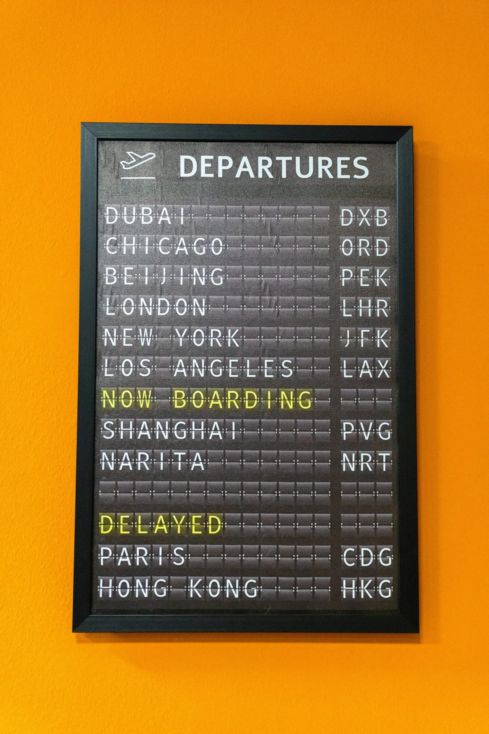 Tableau d'affichage des départs de vols avec destinations comme Dubaï, Chicago, Beijing, Londres, New York, Los Angeles, Shanghai, Narita, Paris, Hong Kong, et leurs codes d'aéroport, sur un mur orange.
