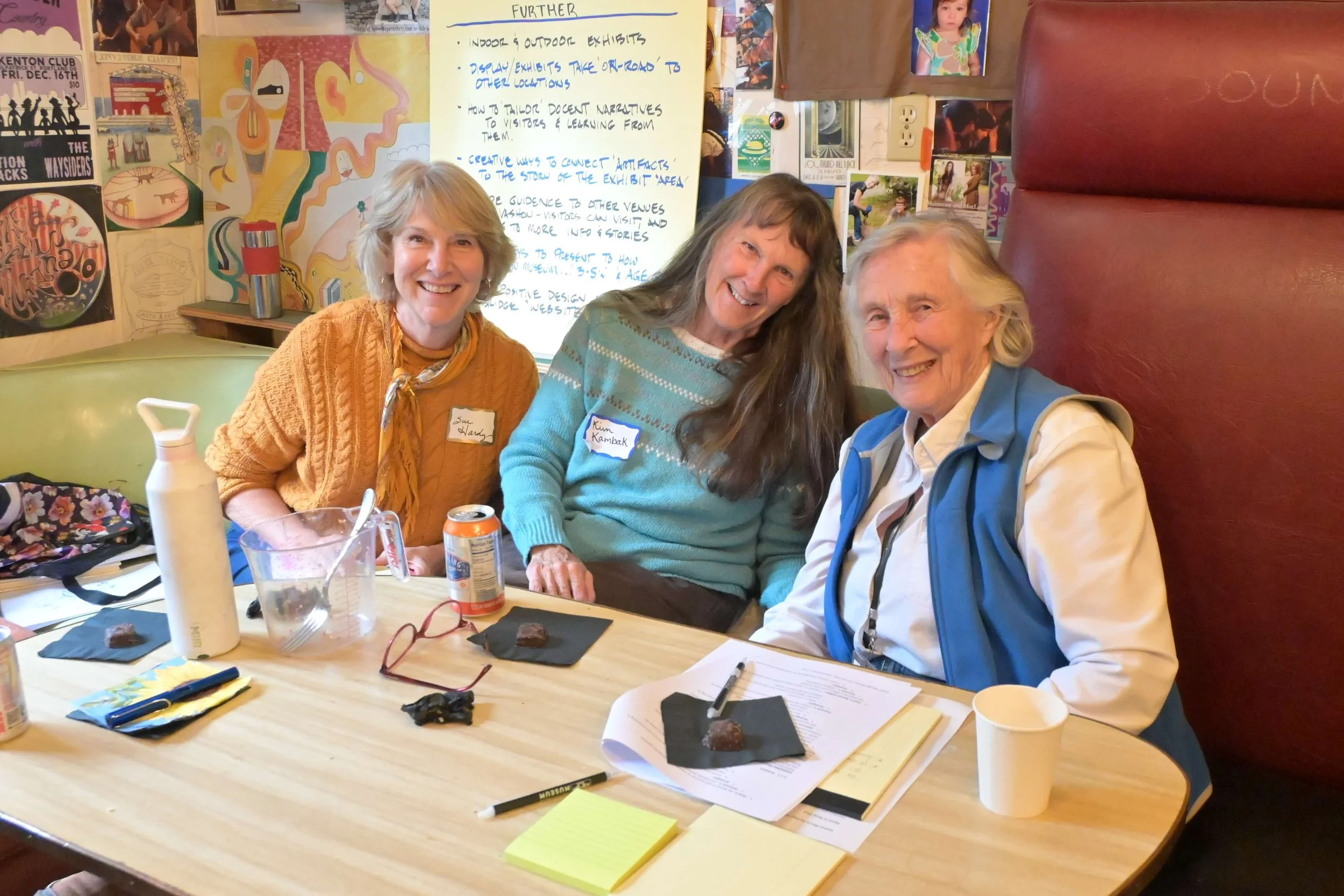 Three women sitting at a table smiling for the camera in a colorful room, with papers, cans, sunglasses, and a water bottle on the table.