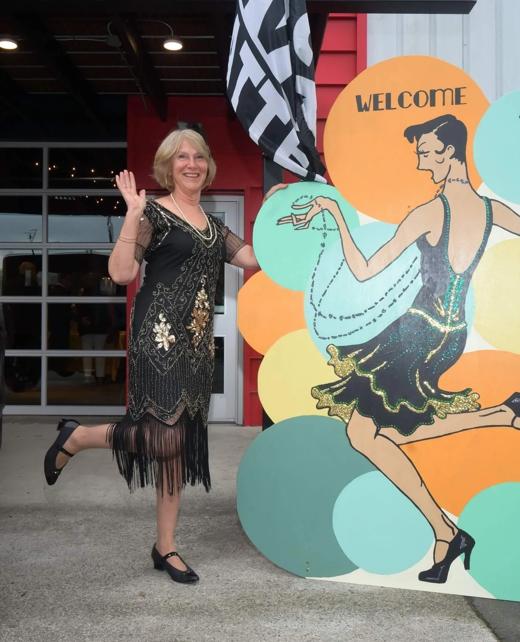 A woman in a black and gold dress with fringe on the bottom, standing outdoors, smiling, and waving next to a colorful cartoon-style sign that reads 'Welcome' with a woman in a dress and high heels.