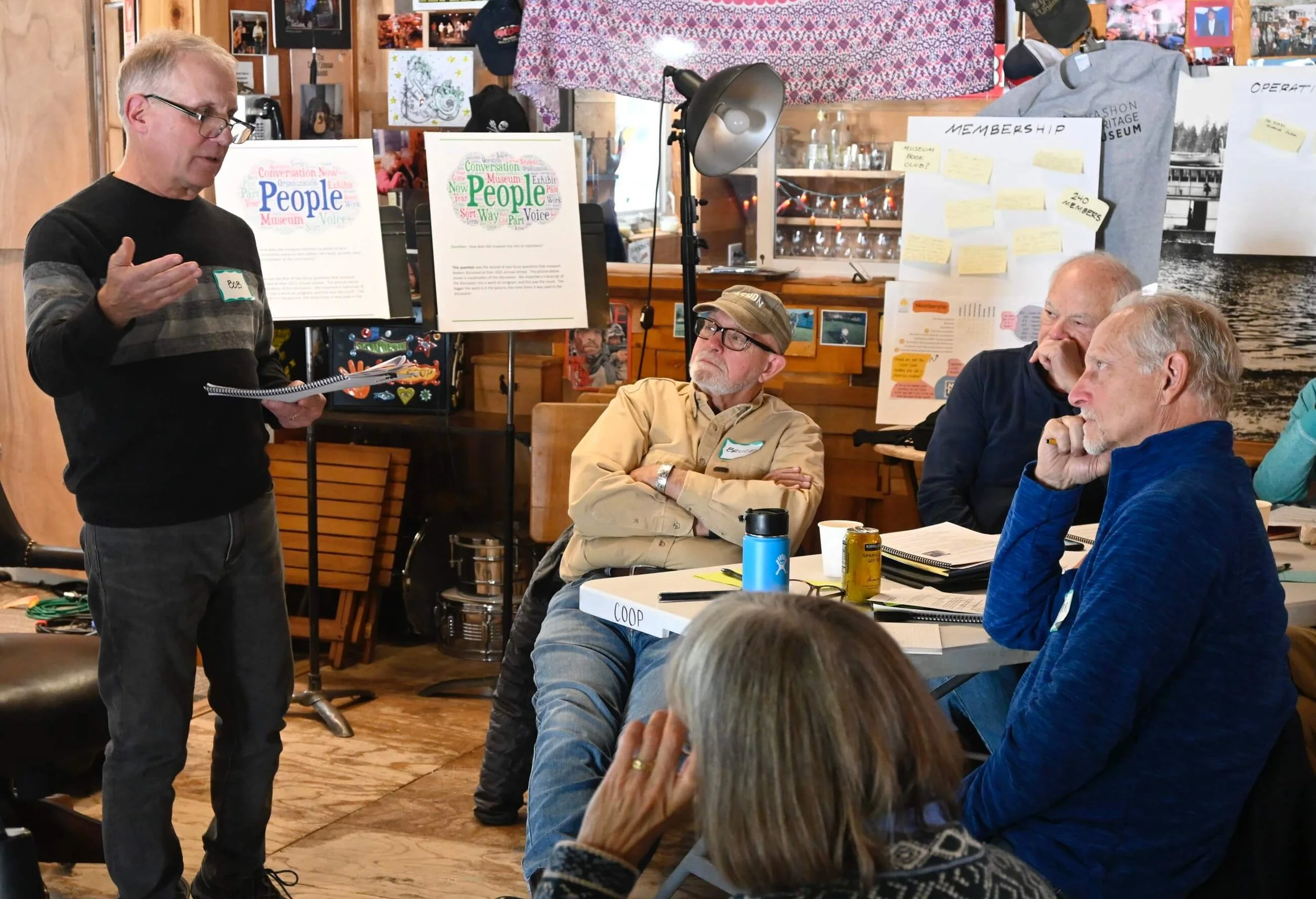 A man standing and speaking to a seated group at a meeting or workshop in a rustic room with wooden walls and various posters and notes displayed.