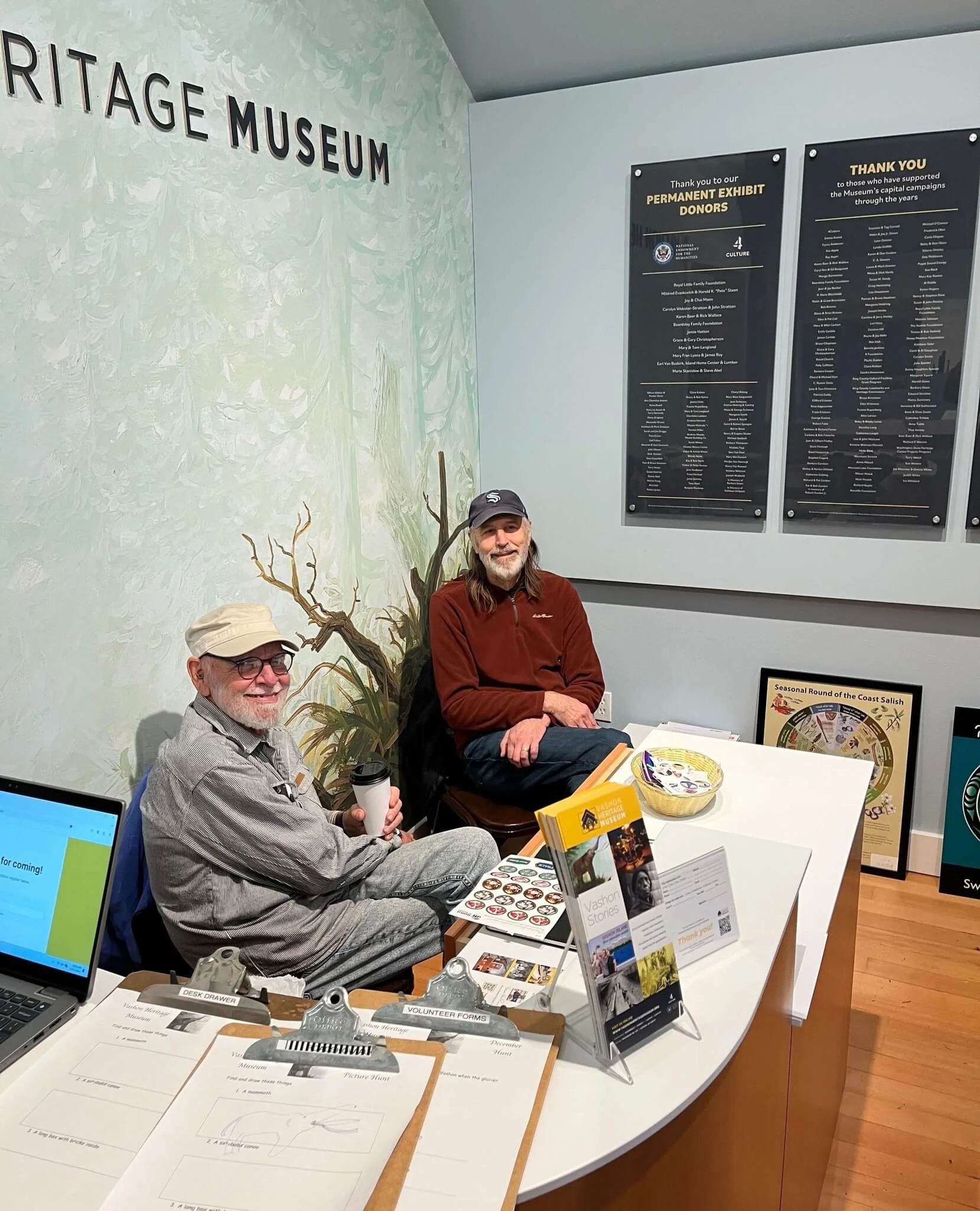 Two men sitting at a table inside a heritage museum exhibit space. The man on the left is wearing glasses, a beige hat, and a gray shirt, holding a travel mug. The man on the right is wearing a dark cap and a red zip-up jacket. There are informational pamphlets, volunteer forms, and a bowl of candy on the table. Behind them, a wall displays information about donors and sponsors, and there are framed posters on the floor leaning against the wall.