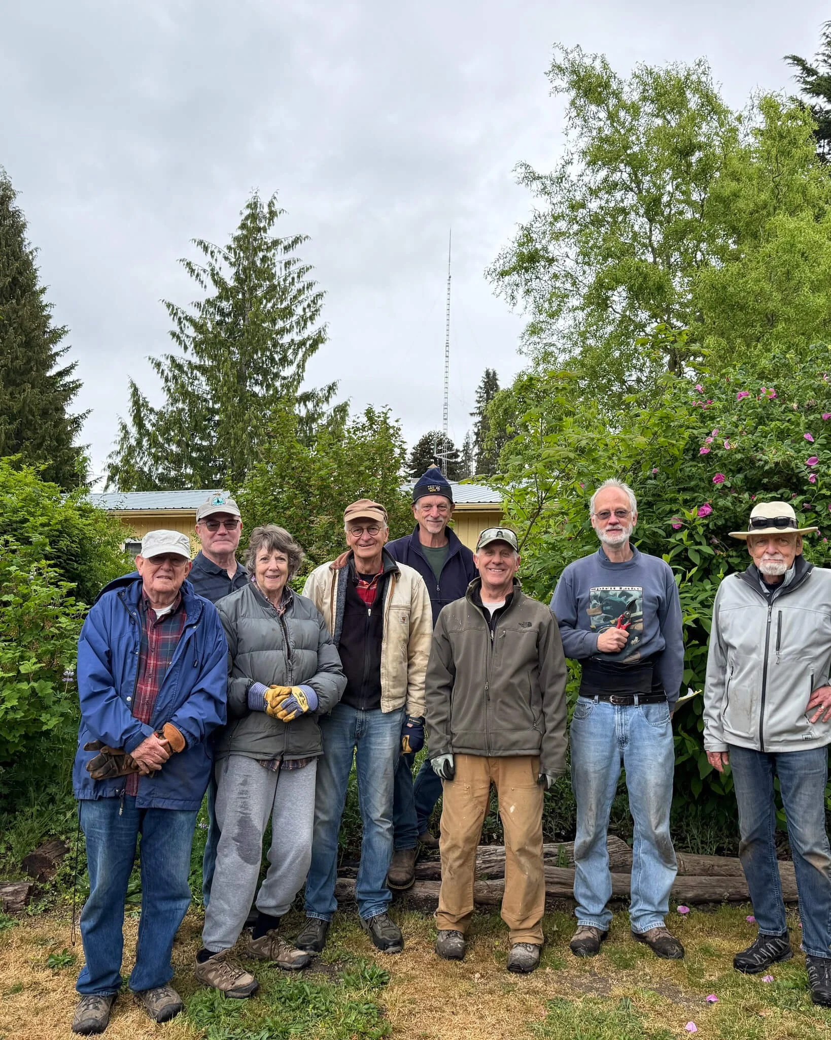 Group of nine adults standing outdoors in front of trees and bushes, dressed in casual outdoor clothing, with some wearing hats and gloves, on a cloudy day.