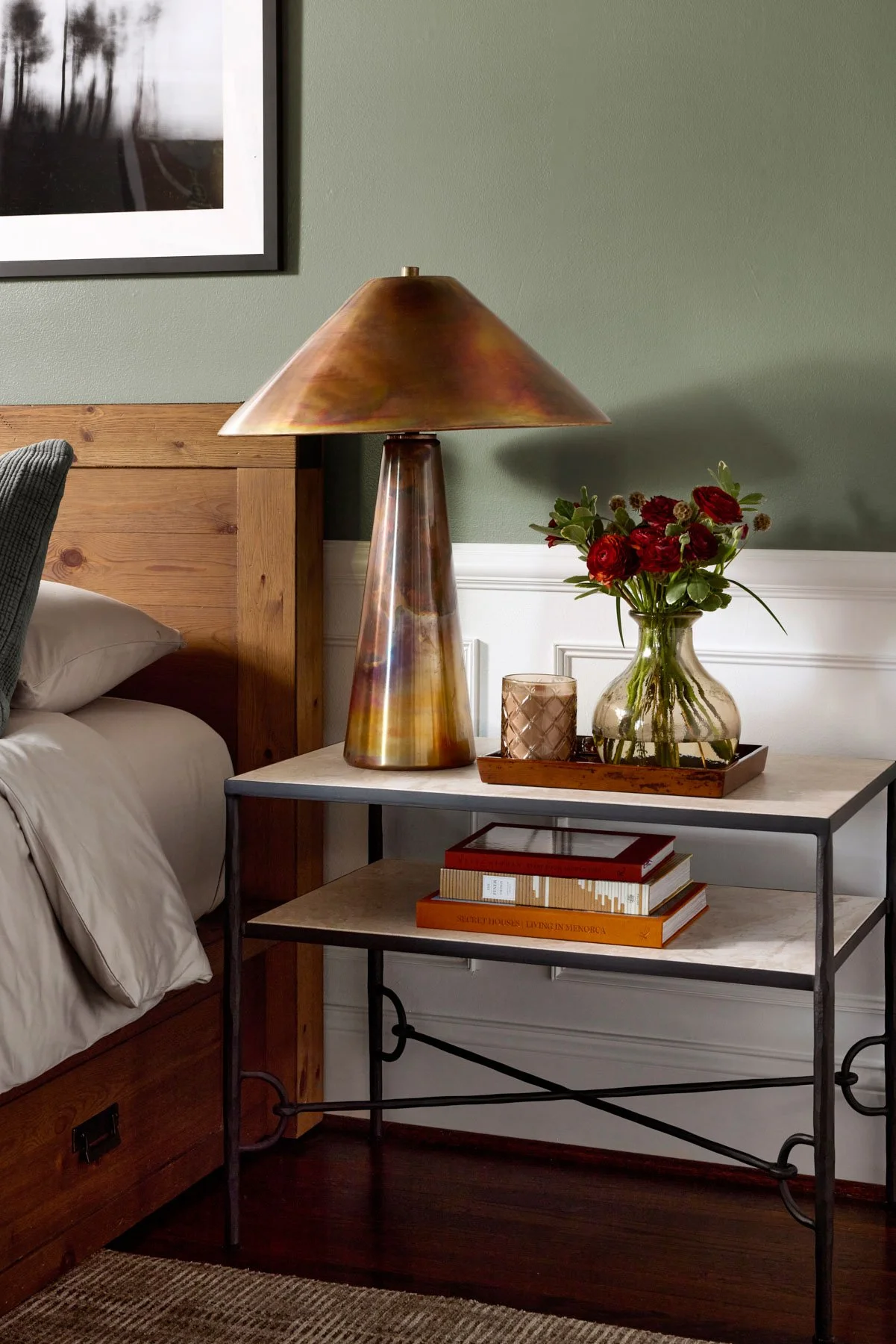A bedroom corner with a nightstand holding a decorative table lamp, a glass vase with red flowers, a candle holder, and books, next to a bed with a wooden headboard and a framed black-and-white photograph on the green wall.