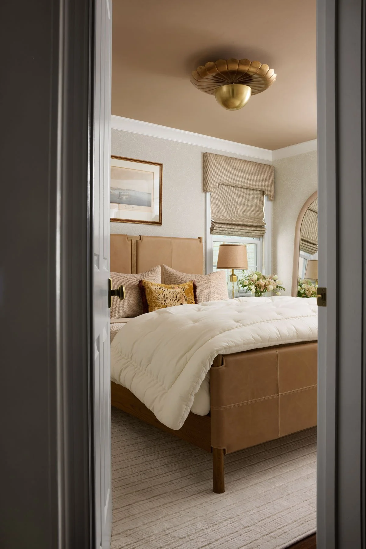 A cozy bedroom viewed through an open door, featuring a bed with a beige upholstered headboard and pillows, a white comforter, a side table with a lamp, a portrait on the wall, and a window with patterned Roman shades and flowers on the windowsill.