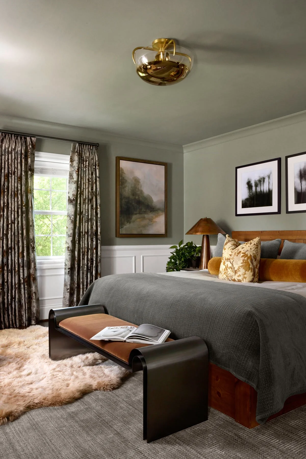 A bedroom with a bed, decorative pillows, a nightstand with a lamp, plants, framed artwork, and a window with floral curtains. There is a black and brown bench at the foot of the bed with an open book. A fluffy rug is on the floor.