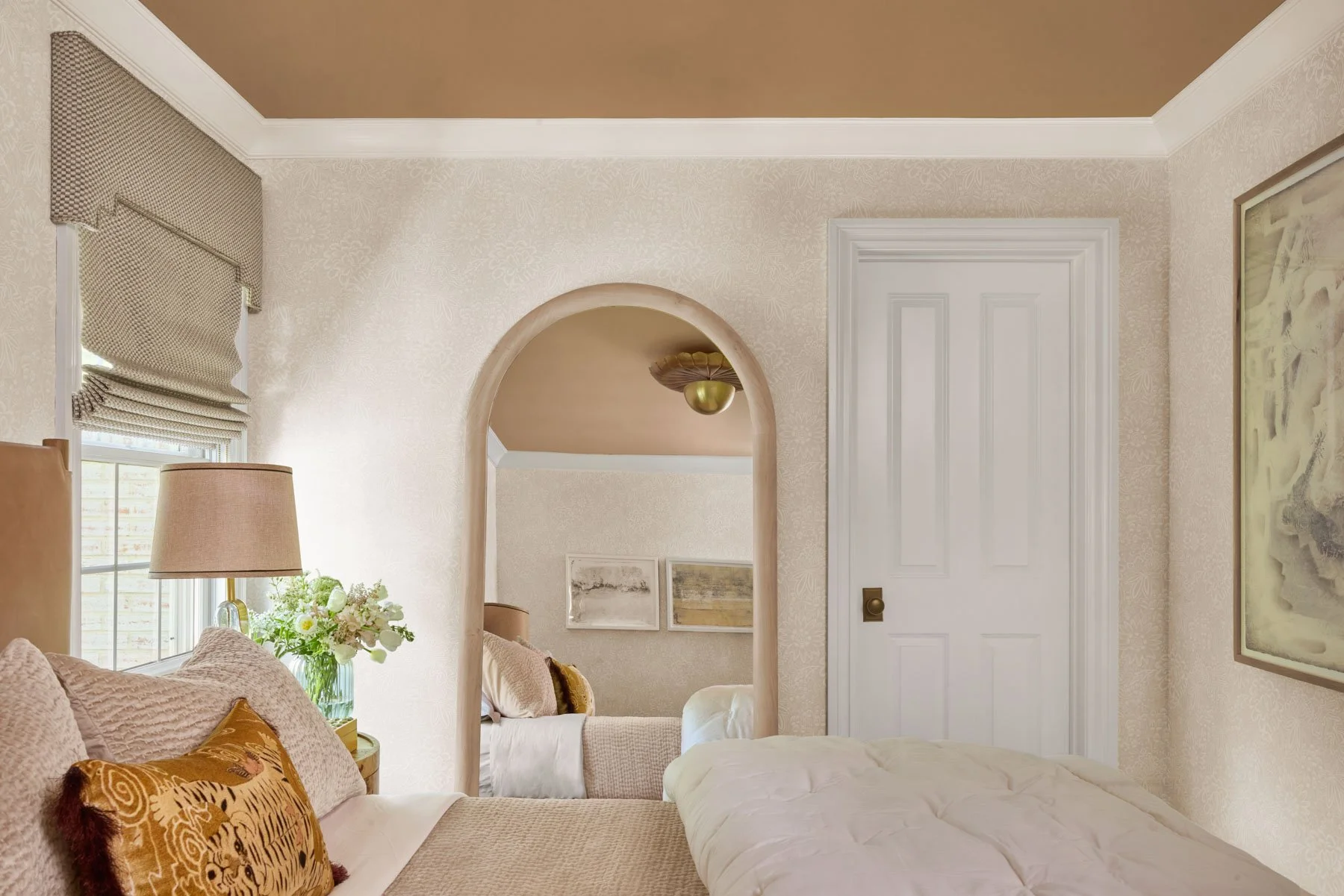 A cozy bedroom with a bed, beige pillows, a green nightstand with a vase of flowers, a lamp, and a window with patterned curtains. An arched doorway leads to another room with framed artwork and a ceiling light fixture.