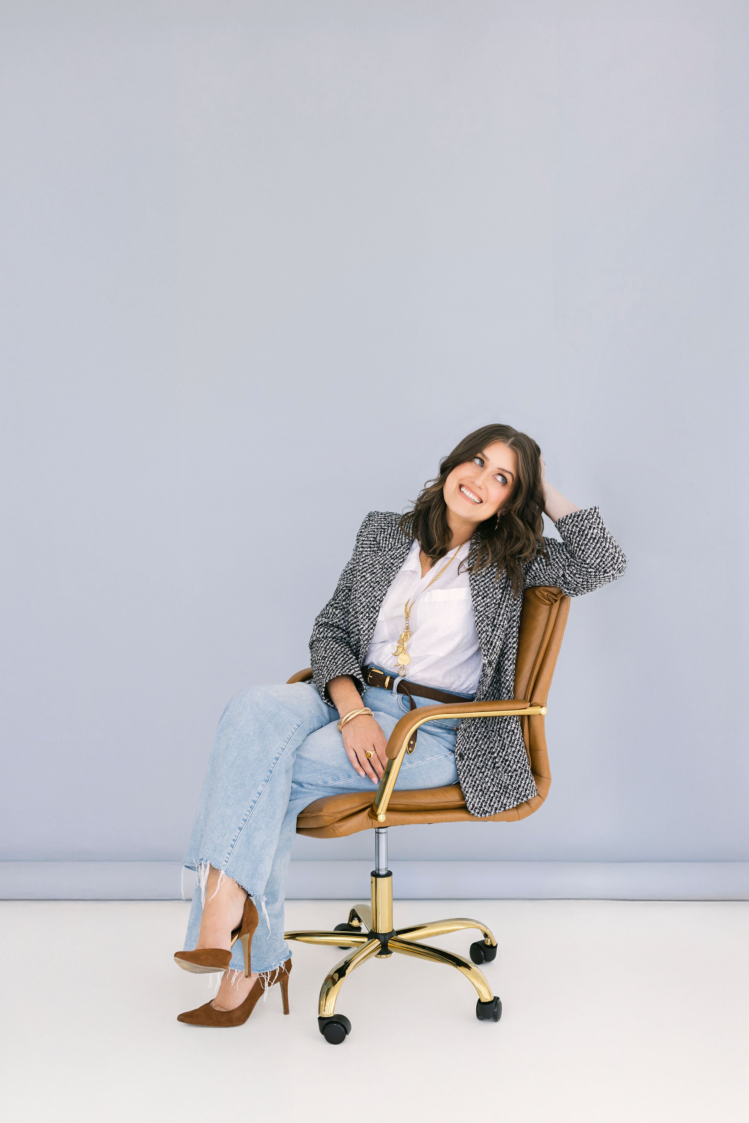 A woman sitting on a brown office chair with a light gray background. She has dark wavy hair, is smiling, and wearing a blazer, white shirt, distressed jeans, and brown high heels.
