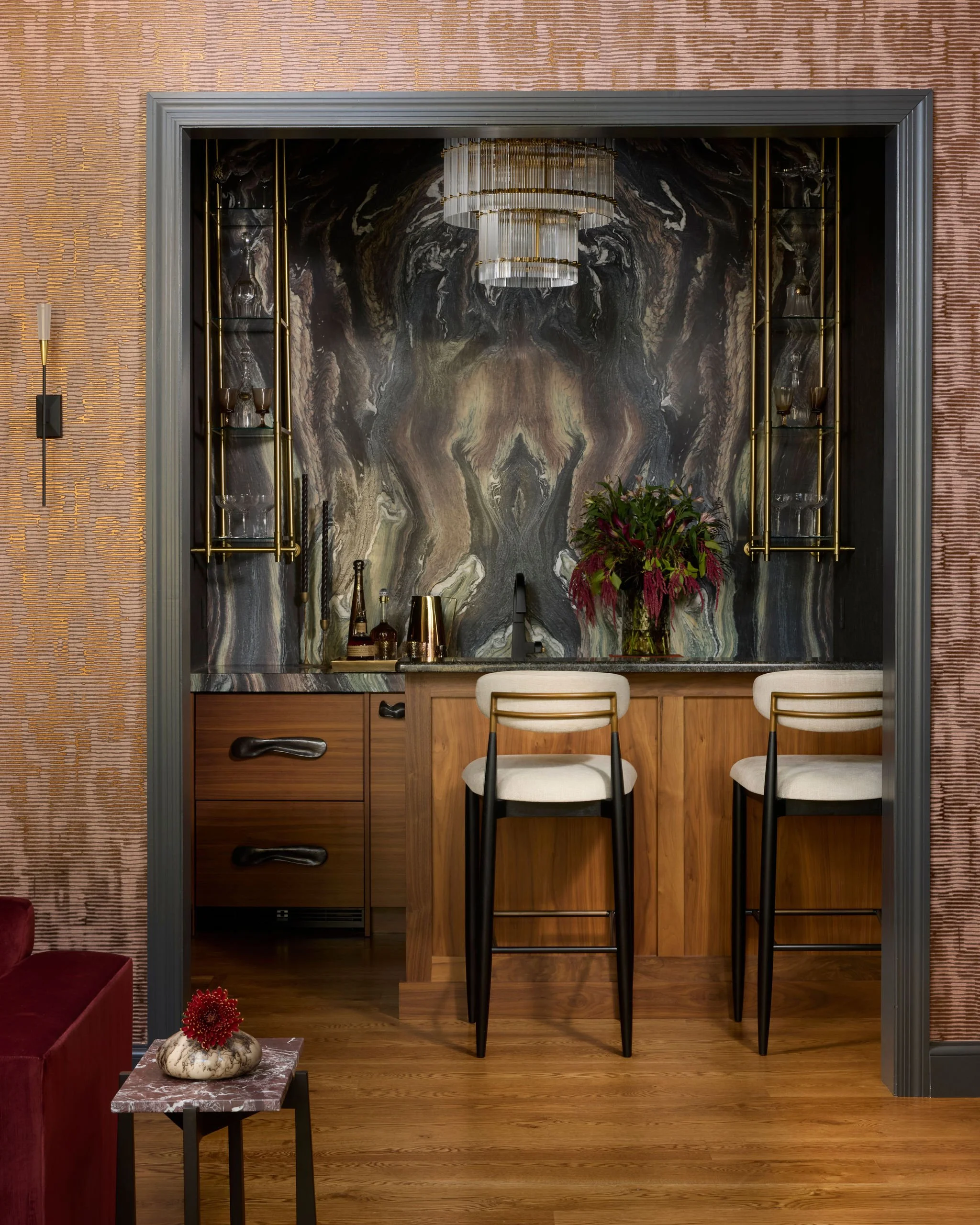 A stylish bar area with a black and gold color scheme, marble background, two beige barstools with black frames, a wooden counter, a large floral arrangement, and decorative glass shelving.