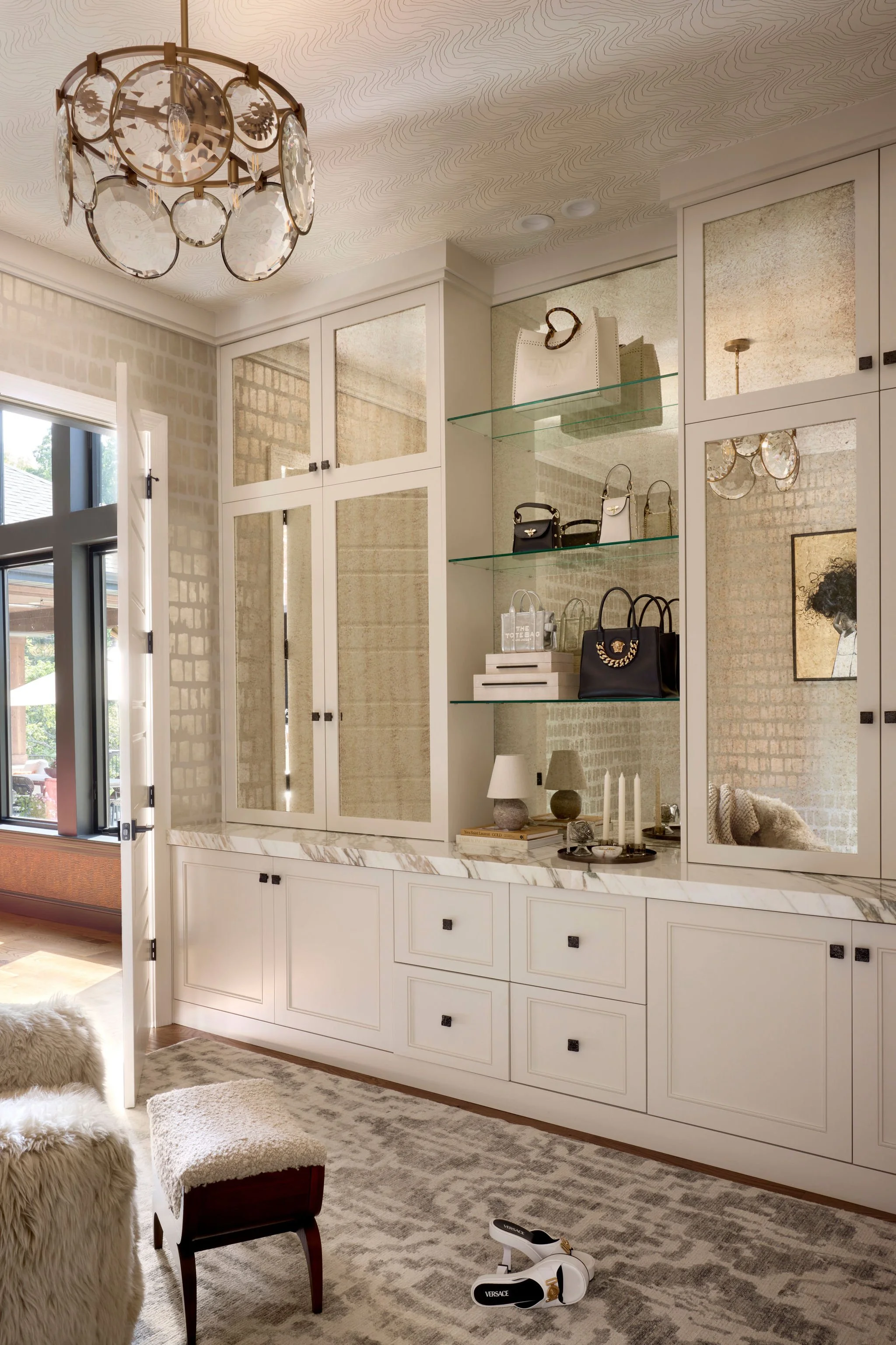 Living room with built-in white cabinet and glass shelves displaying handbags, art, and accessories, light-colored patterned rug, plush seating, and a decorative chandelier.