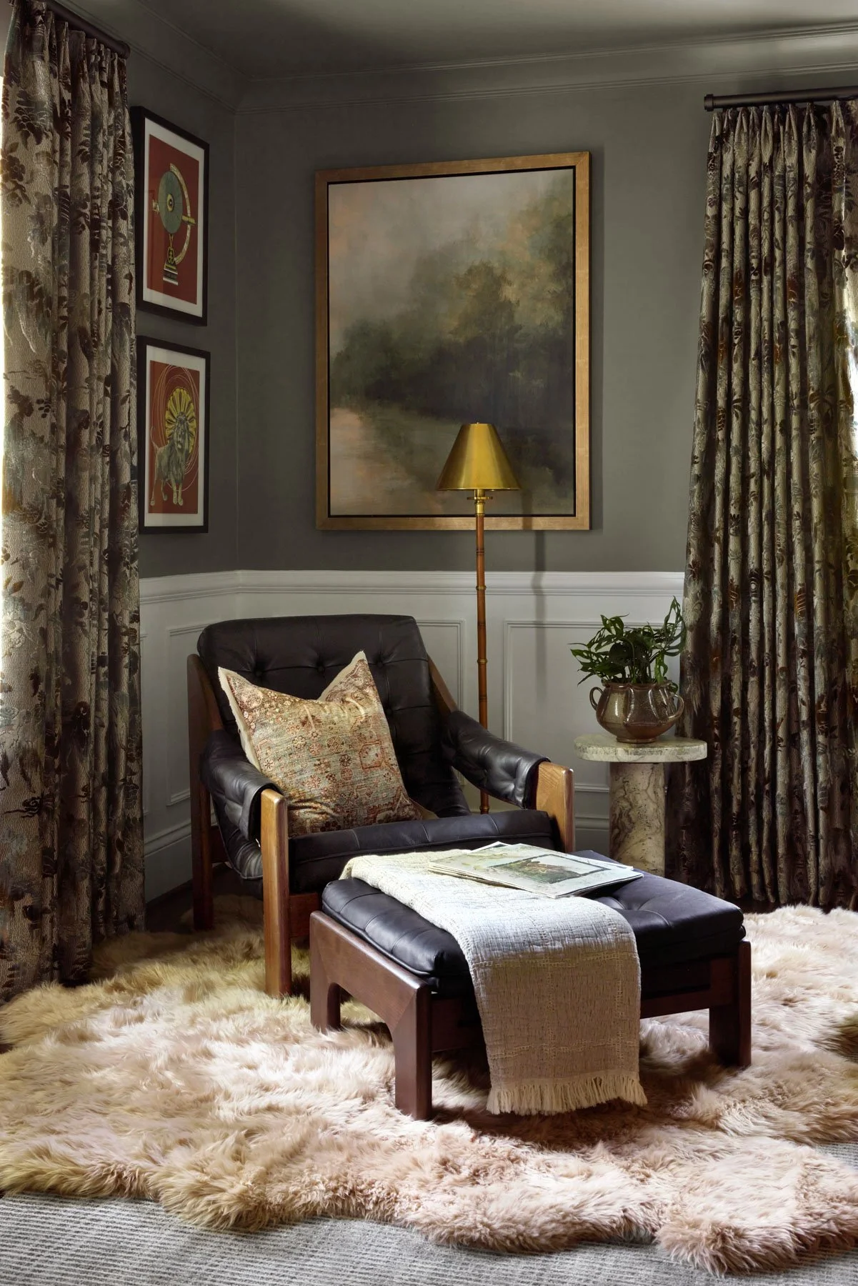 A cozy living room corner with a black leather armchair, a matching ottoman with a beige throw, and a fluffy cream-colored area rug. There are patterned curtains on both sides, a large framed landscape painting, a tall brass floor lamp, and a small m