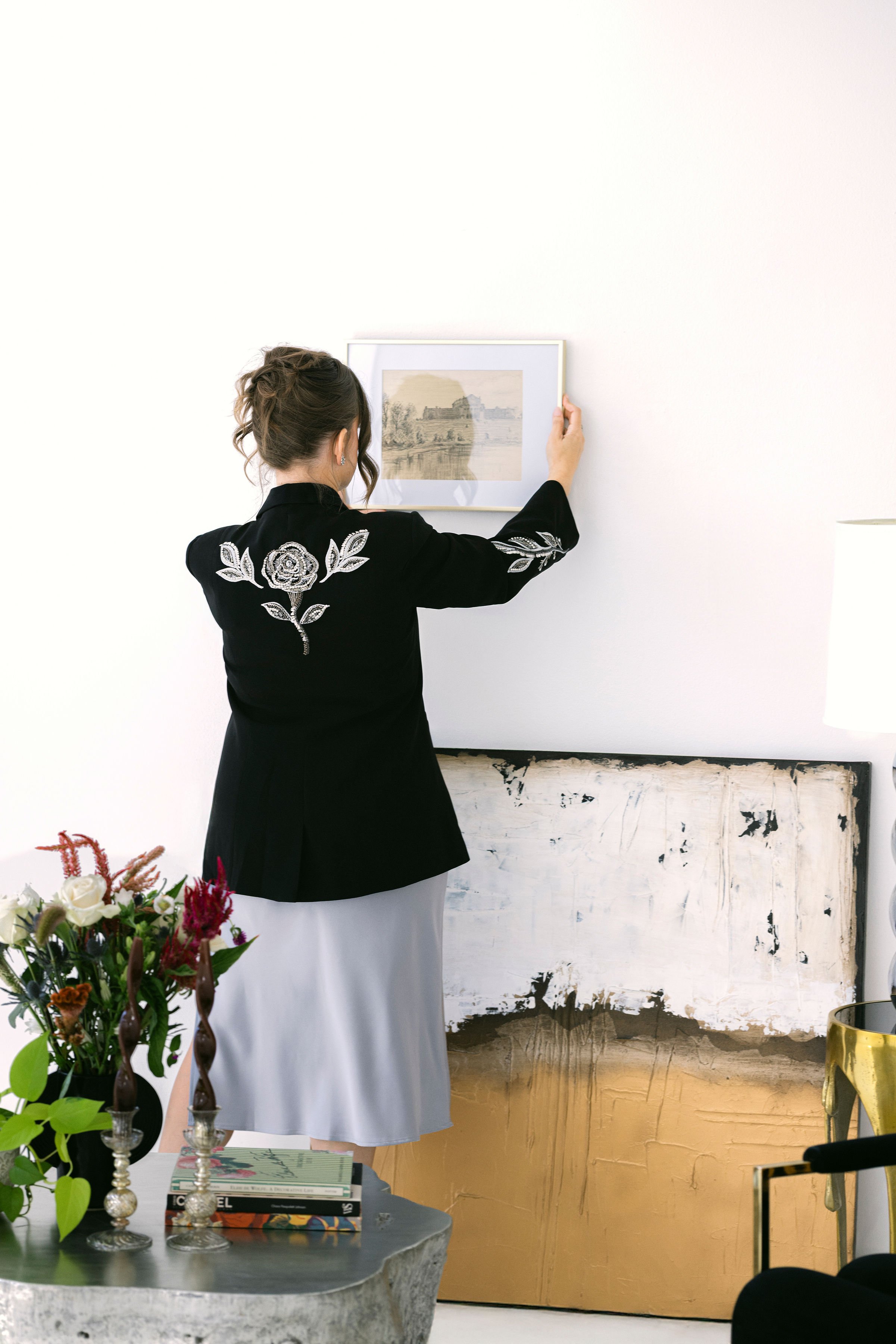 A woman wearing a black blazer with white floral embroidery on the back and sleeves, holding a framed picture on a white wall in an art gallery or home. There are vases with flowers and a stack of books on a table in the foreground, and an abstract painting leaning against the wall.
